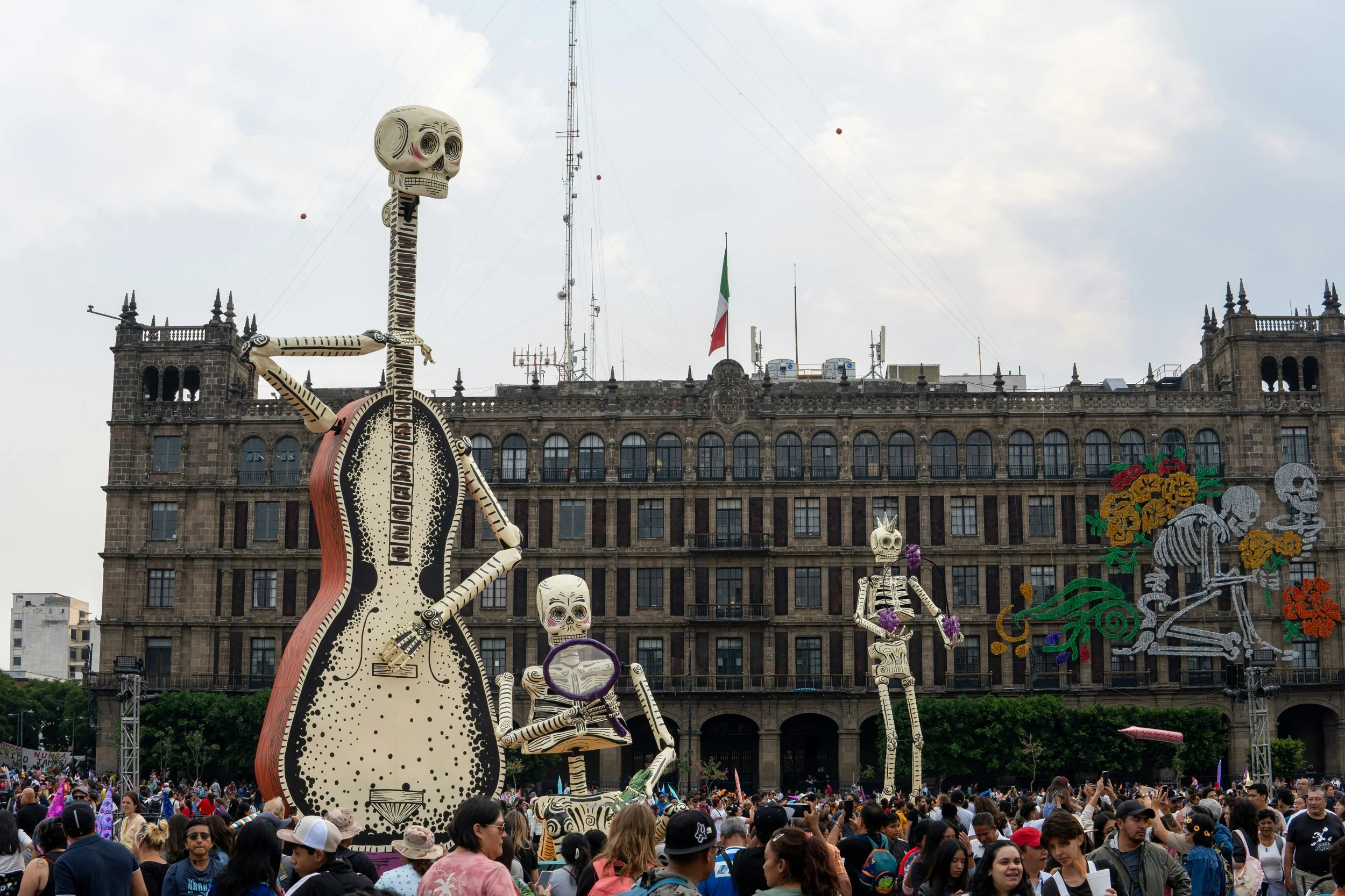 Large skeleton figures, including a giant one playing guitar, at a crowded festival with many people and decorative lights against a historic building with Spanish flag in Mexico City.