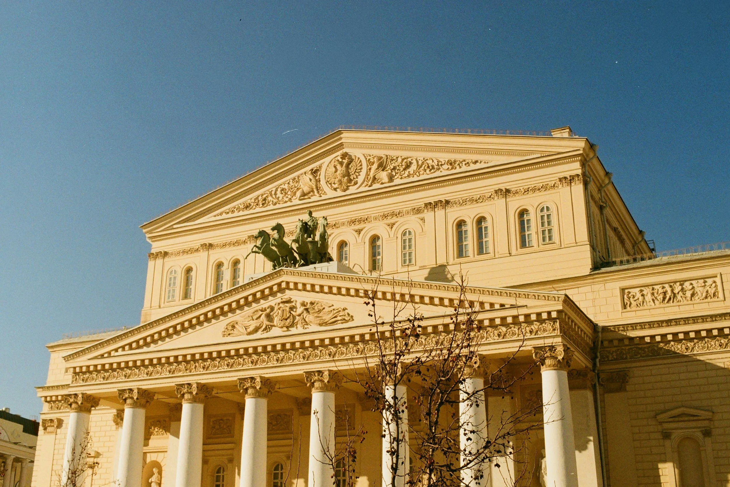 The neoclassical building features a row of white columns supporting a decorated pediment with sculptures, and a statue of a chariot with horses on top. The sky is clear and blue.
