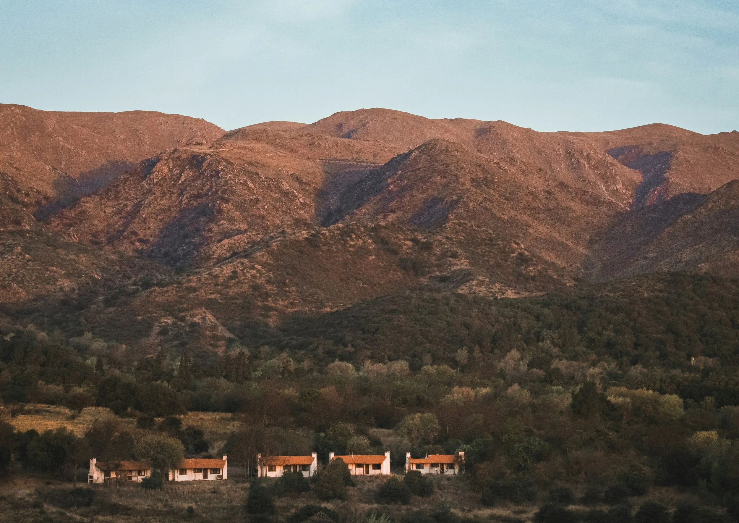 A row of small houses with red roofs and white walls at the base of brown, rolling hills.