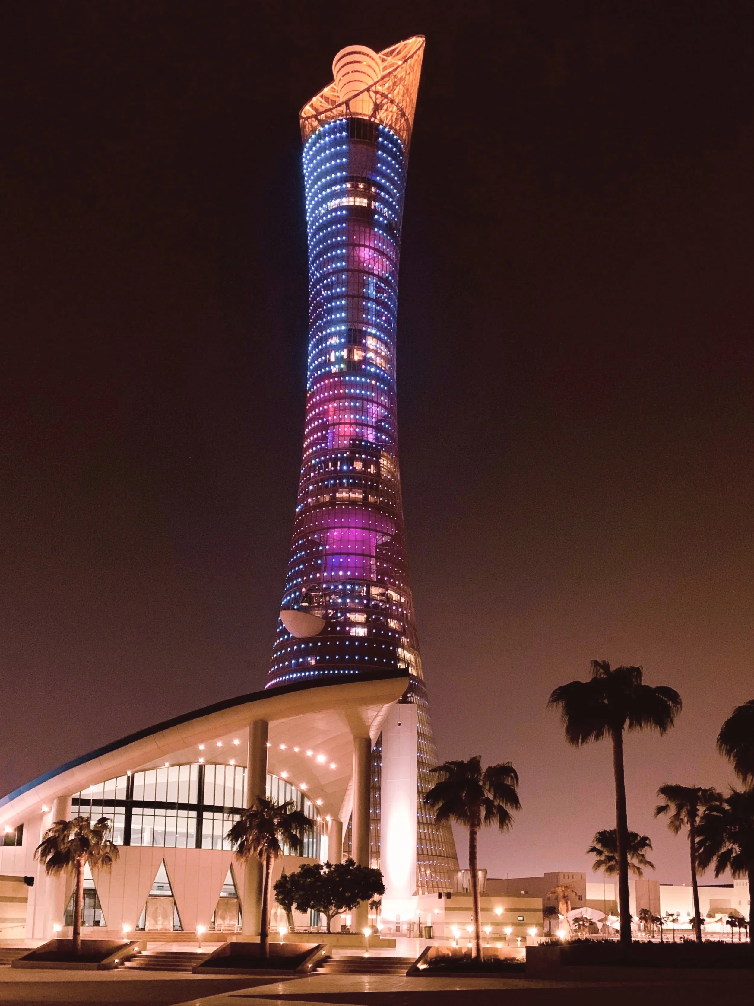 Night view of a tall, illuminated skyscraper with colorful lights and a modern architectural design, with palm trees in the foreground.
