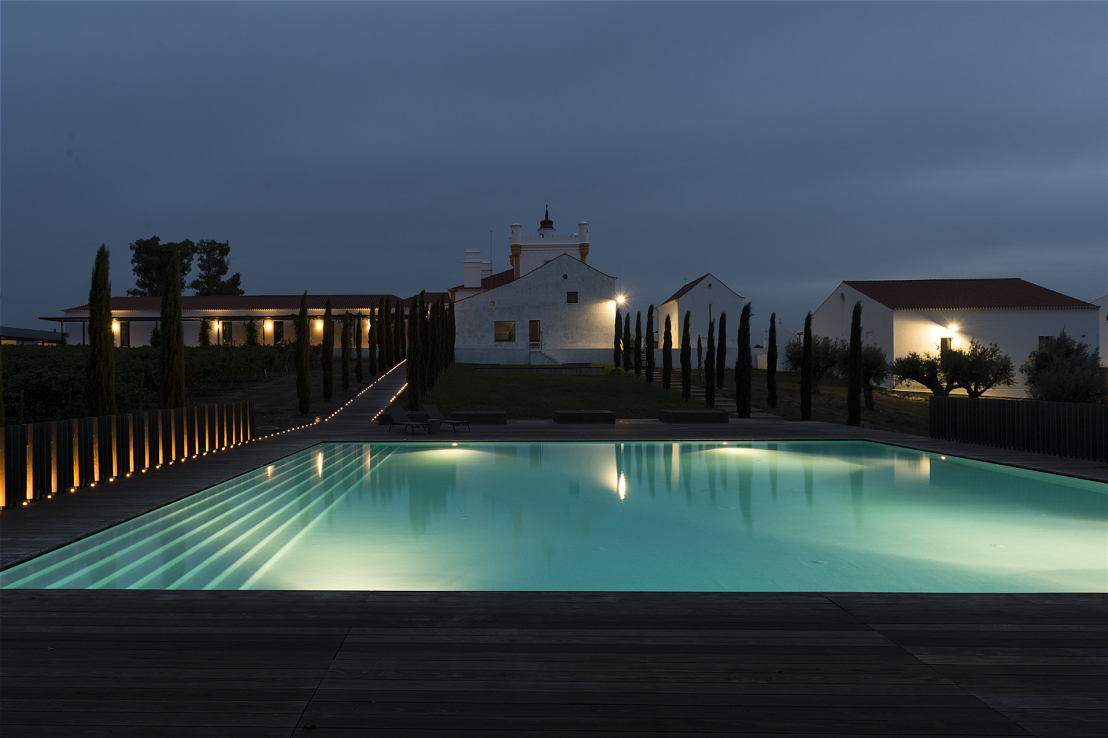 A lit swimming pool with steps leading into the water, surrounded by a wooden deck, with several buildings and tall trees in the background under a cloudy evening sky.