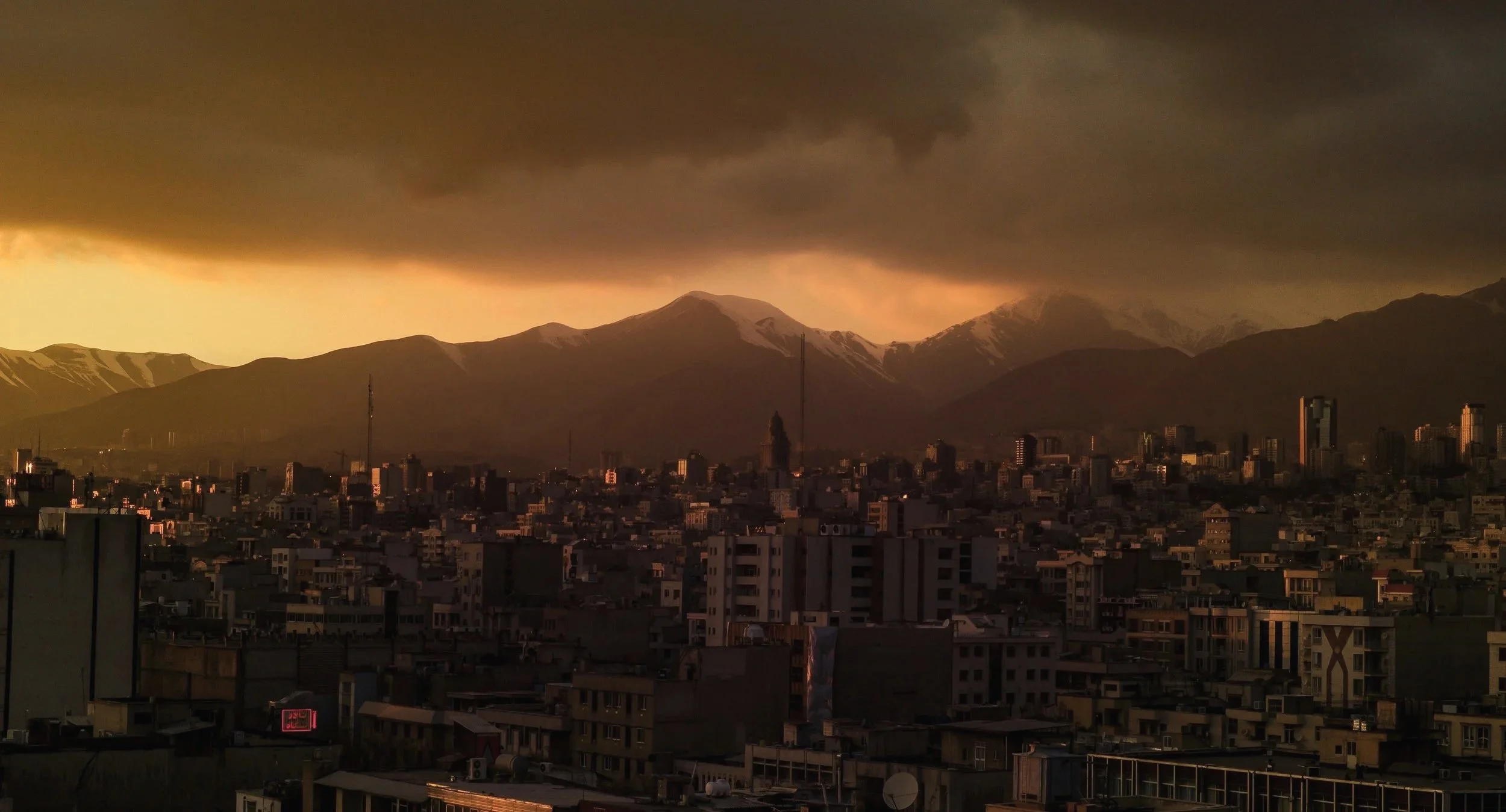 Cityscape with tall buildings in the foreground and snow-capped mountains under a dark, stormy sky at sunset.