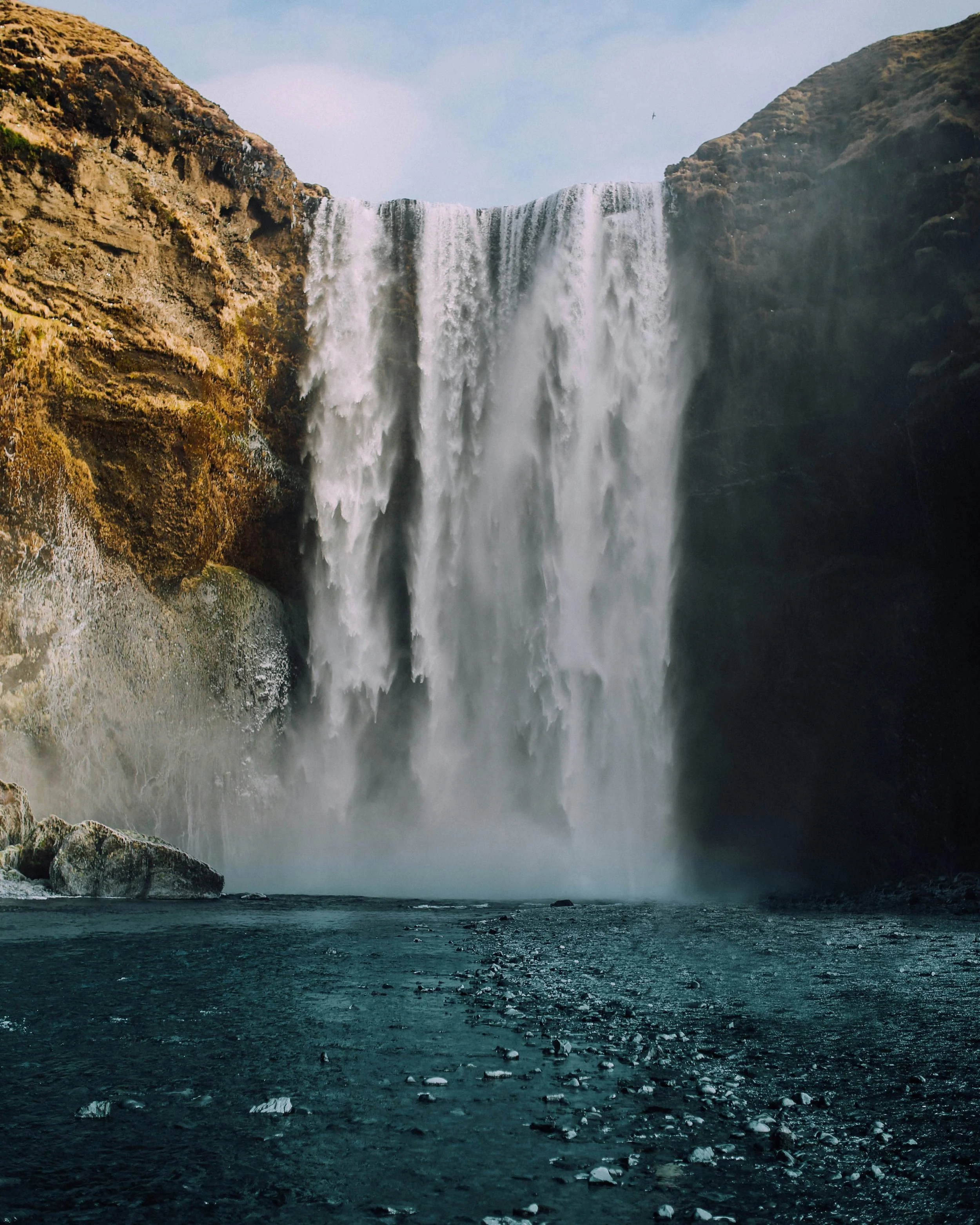 A large waterfall cascading down a rocky cliff into a deep pool of water below, surrounded by rugged terrain.