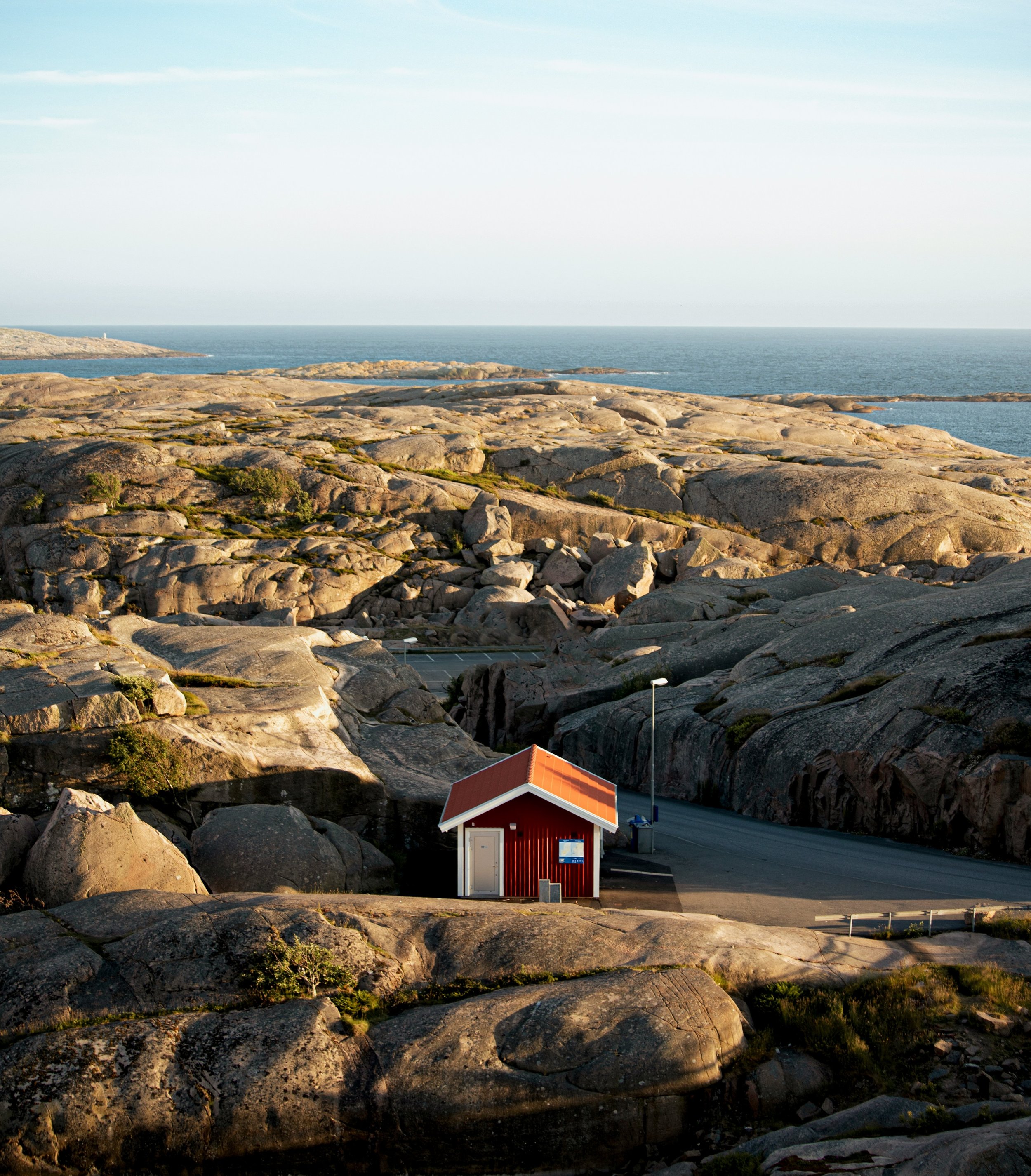 A small red house with a orange roof in a rocky coastal landscape, with the ocean and clear sky in the background.