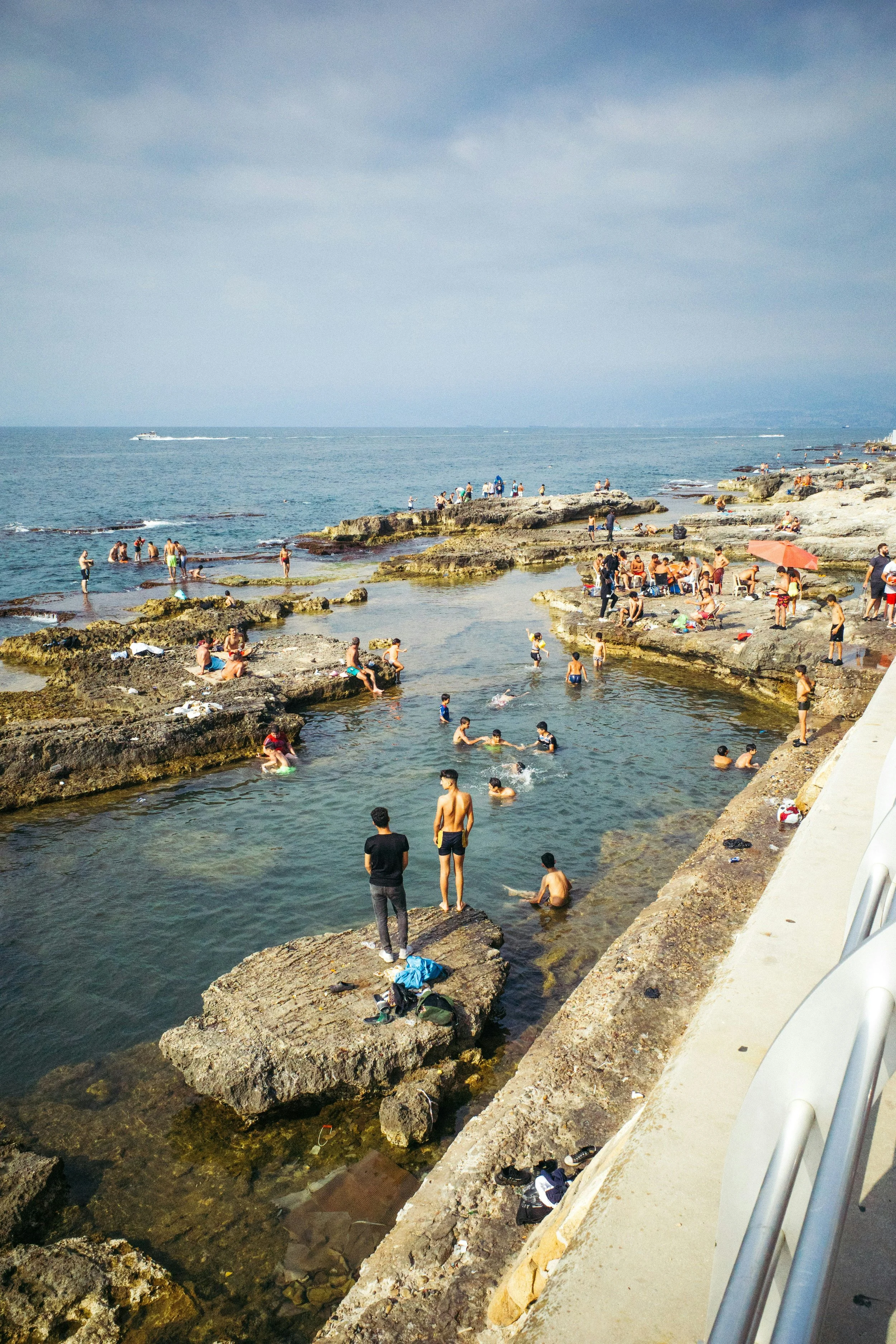 People swimming and relaxing on rocks and in a small pool at a beach with an ocean in the background under a cloudy sky.