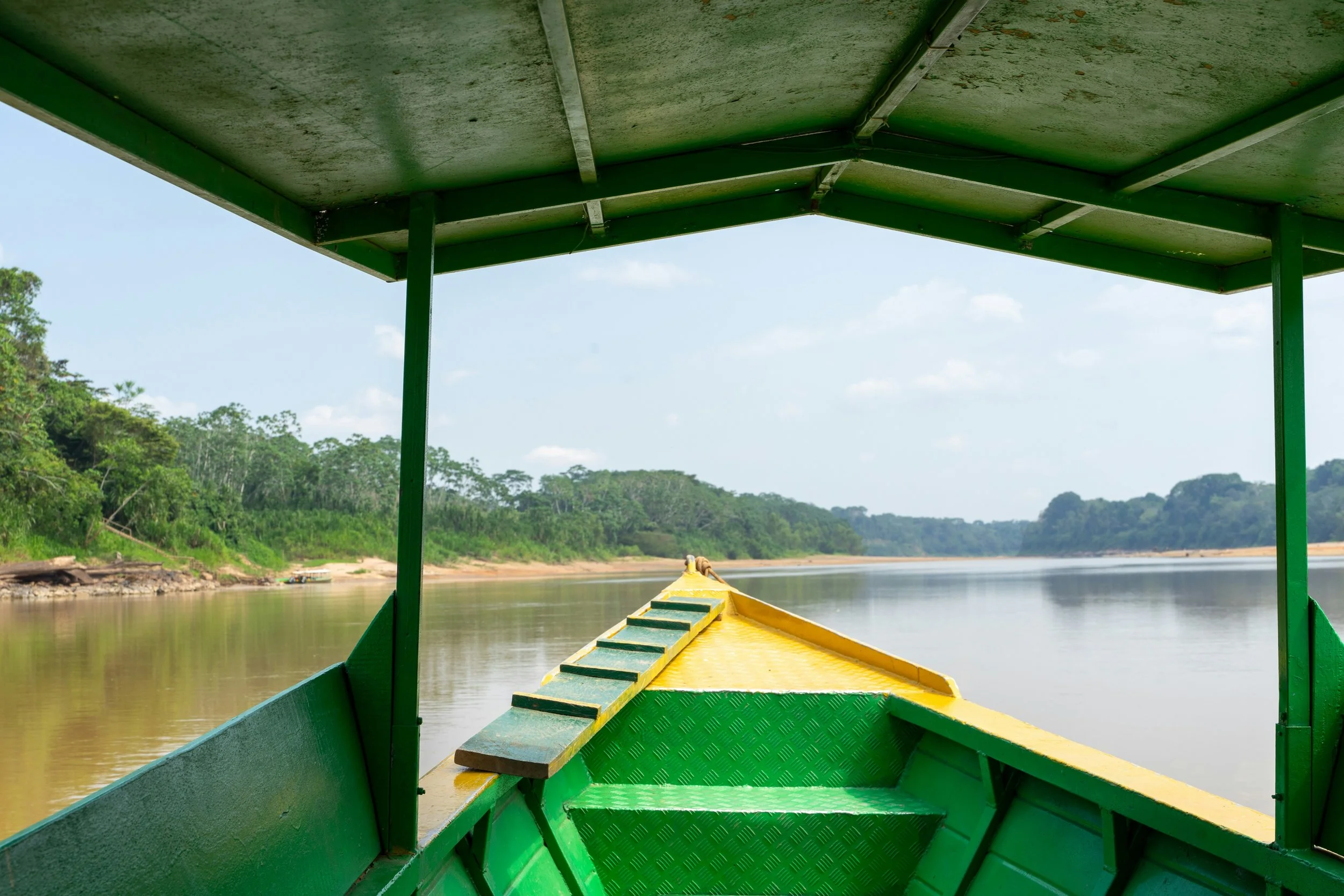 View from a boat on a river showing the boat's green canopy and yellow bow, with calm water and lush green trees along the riverbank under a partly cloudy sky.
