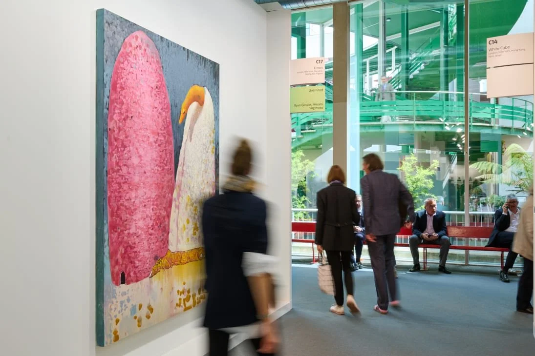 A wildlife ranger in a tan uniform and black vest with patches and text, standing in a forest with dense green foliage, holding a wooden walking stick and gesturing with his hand.