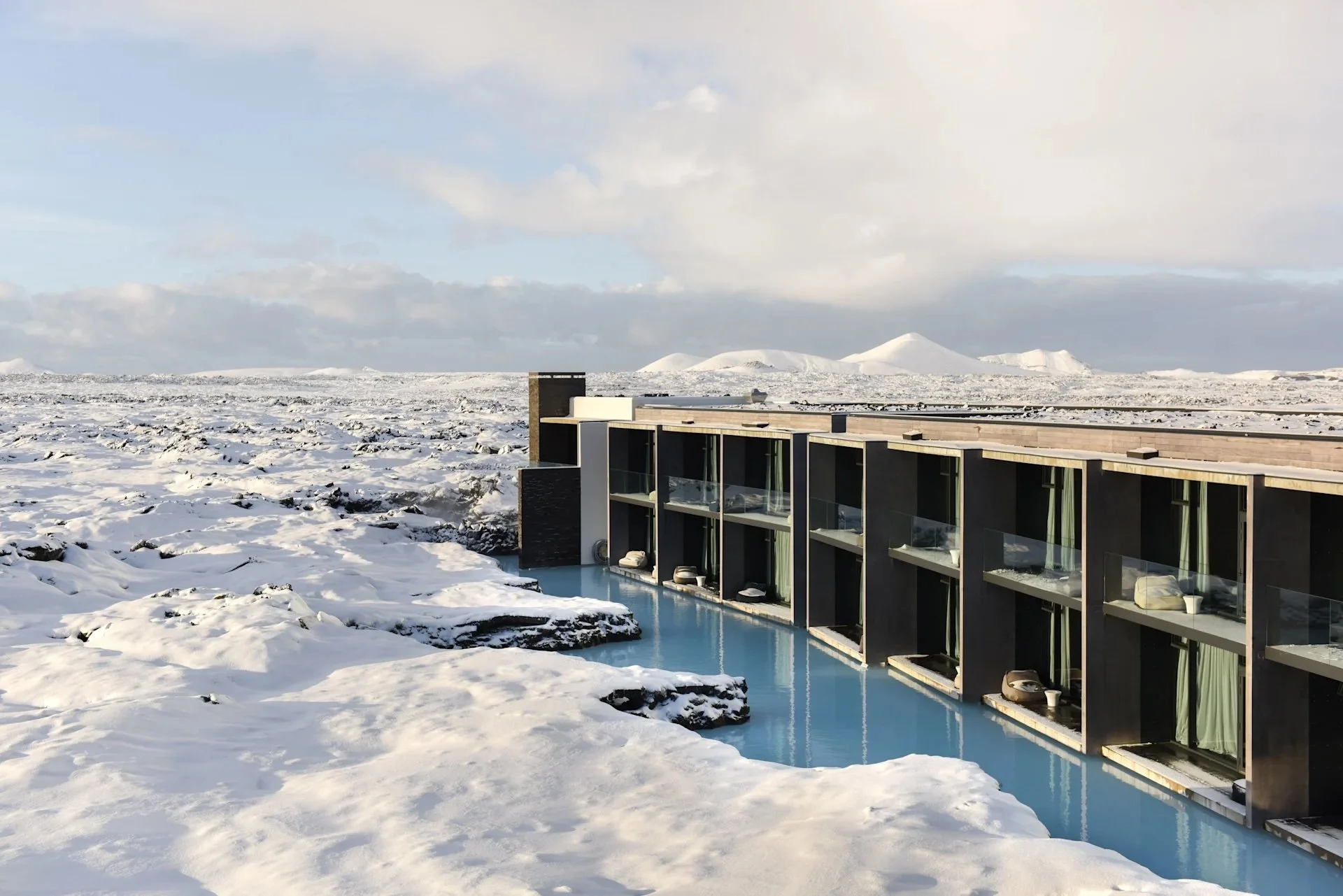 Luxury hotel with glass-fronted rooms overlooking a snowy landscape and a small swimming pool, in a remote icy region with snow-covered mountains in the background.
