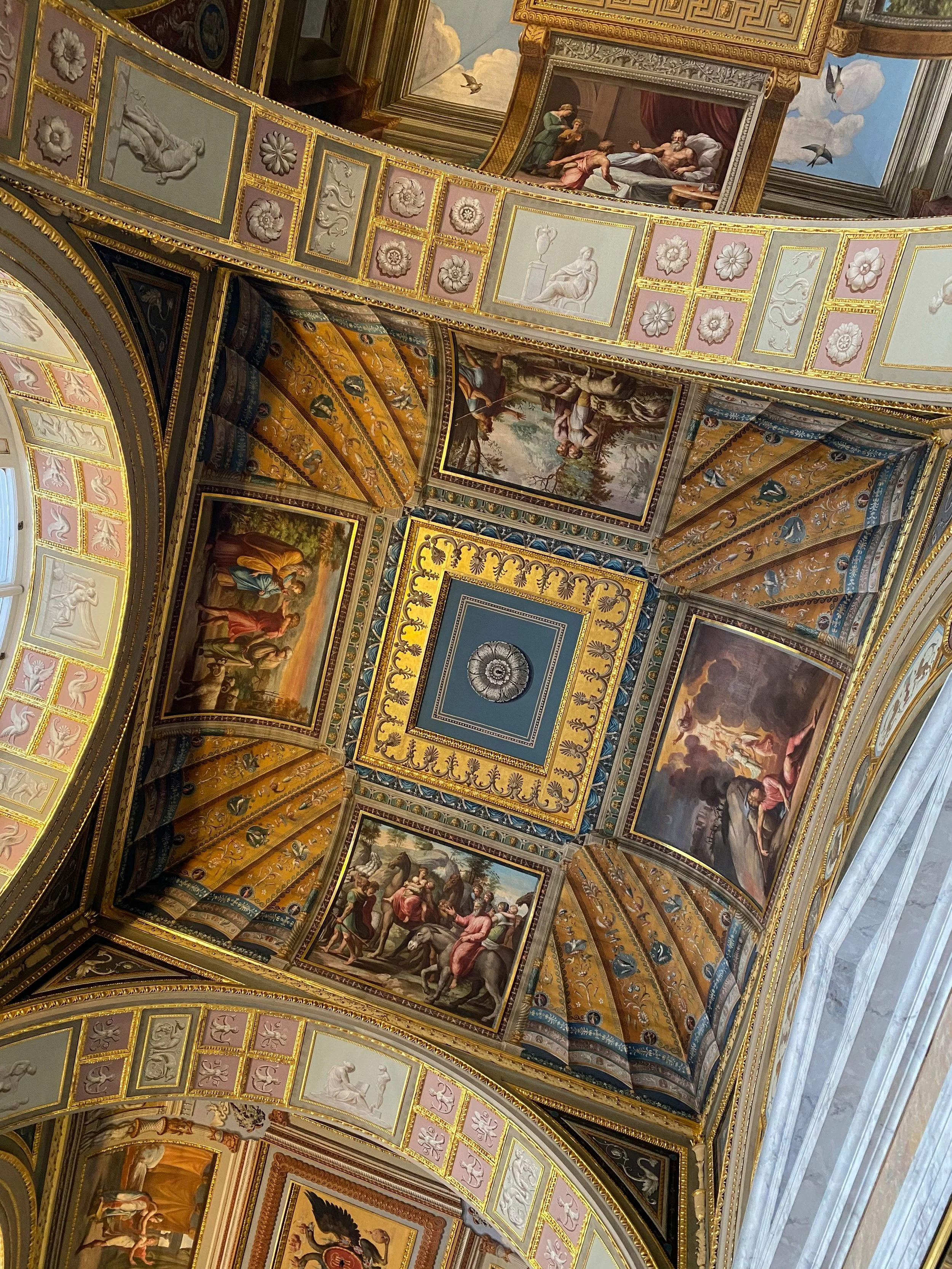 Intricate ceiling with elaborate paintings and gold accents, featuring biblical or classical scenes, floral and geometric motifs, and artistic framing.