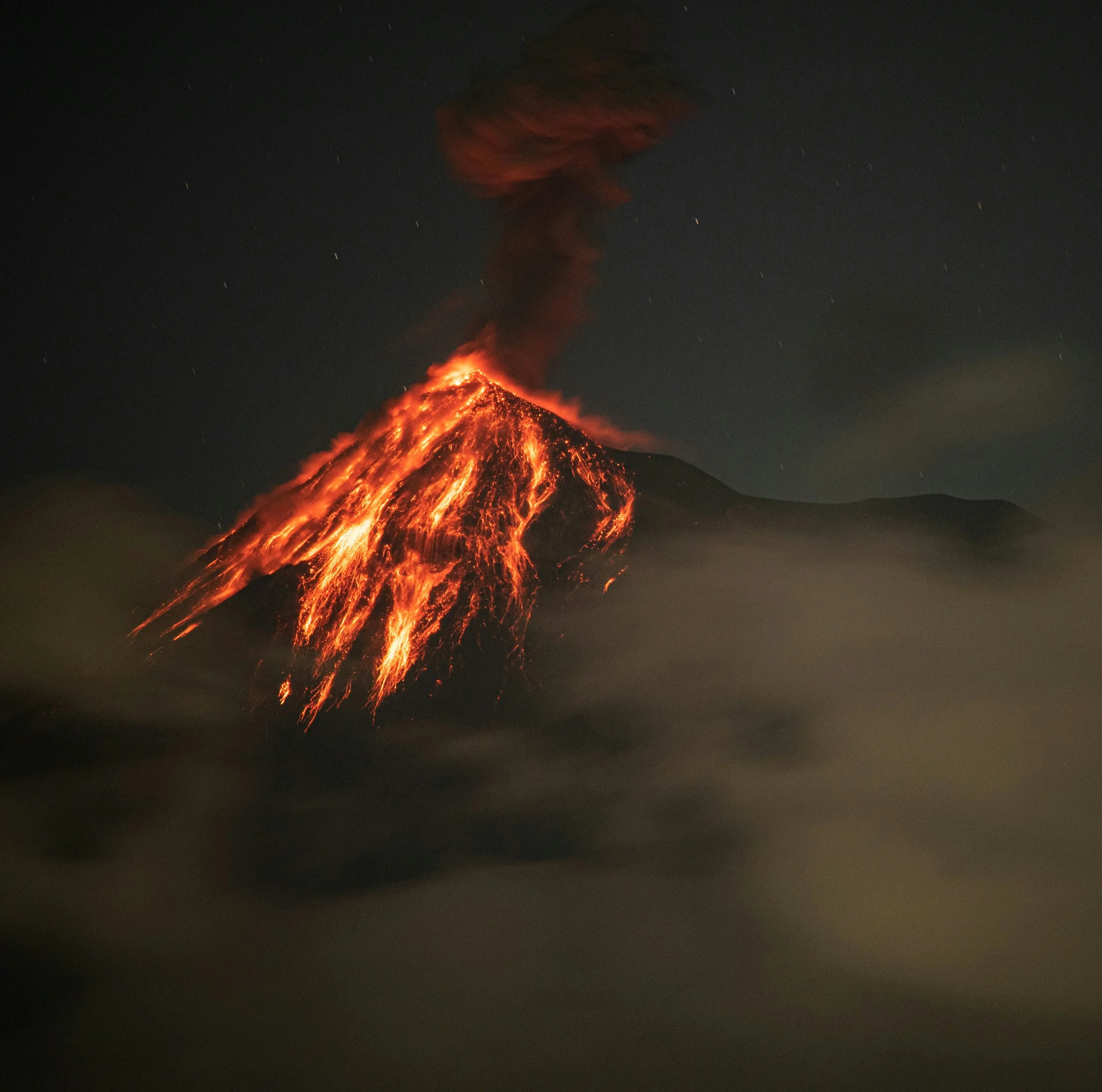 Active volcano erupting at night, with lava flowing down the sides and dark smoke rising from the crater.