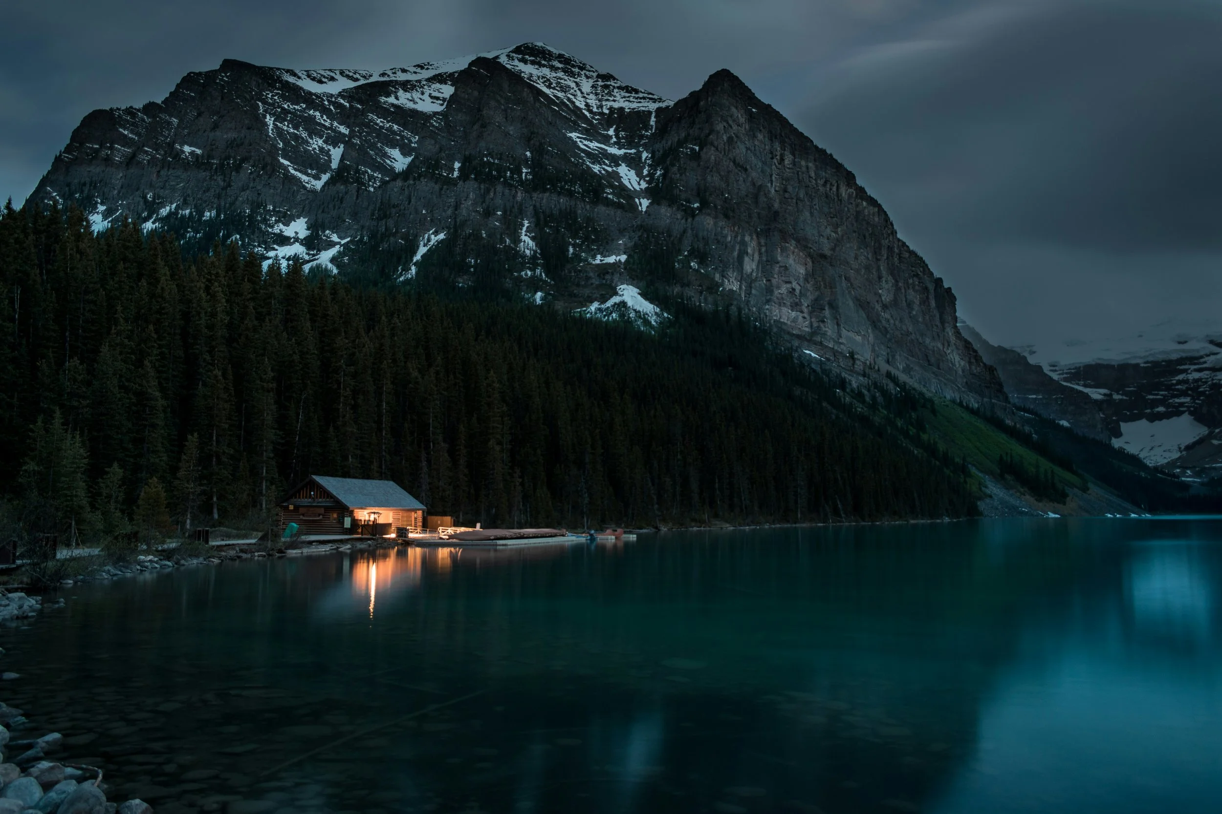 Nighttime view of a mountain lake with a small house emitting warm light, surrounded by dense forest, with snow-capped mountains in the background.