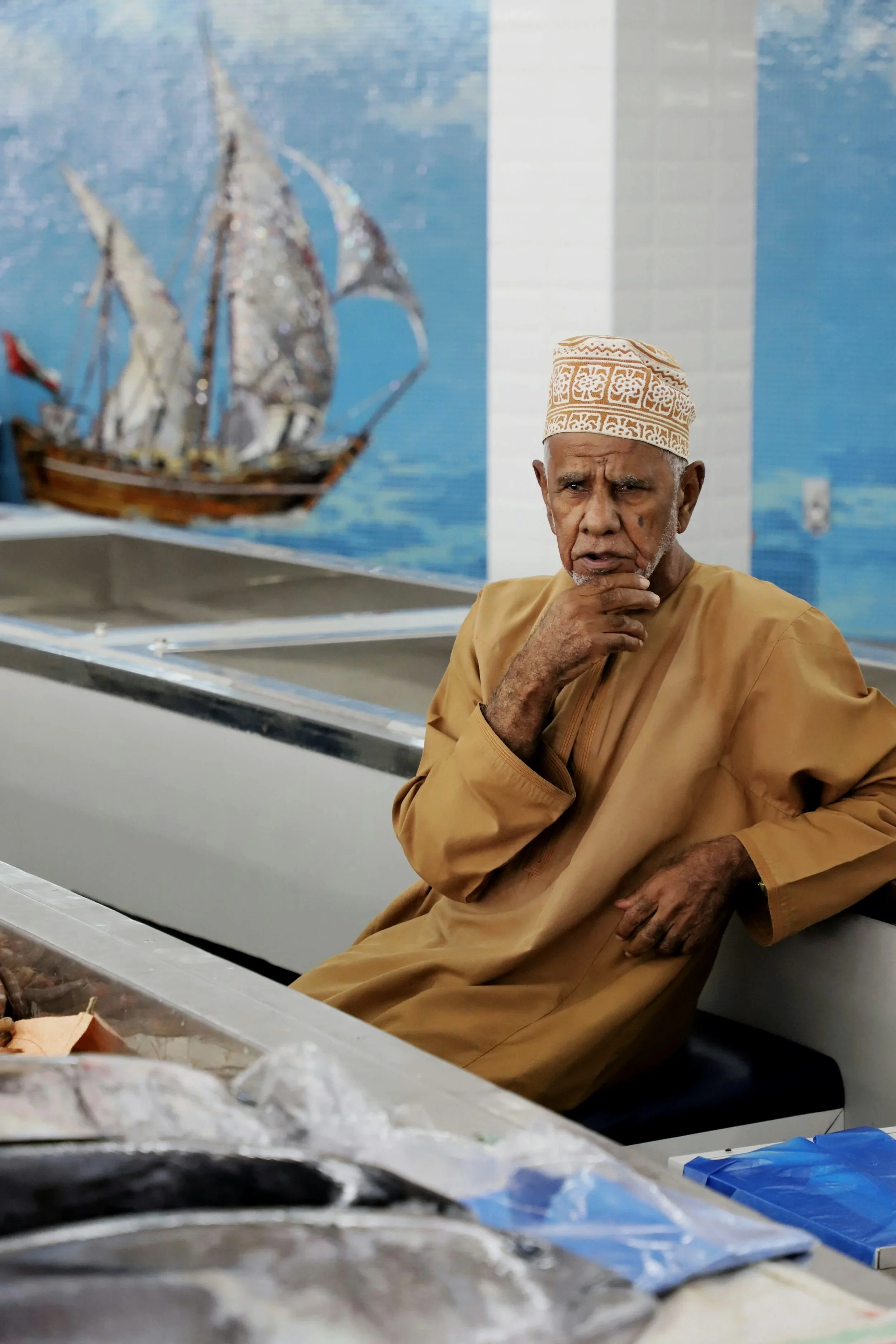 An elderly man wearing traditional clothing and a patterned cap sits in a food shop, with a mural of a sailing ship on the wall behind him.