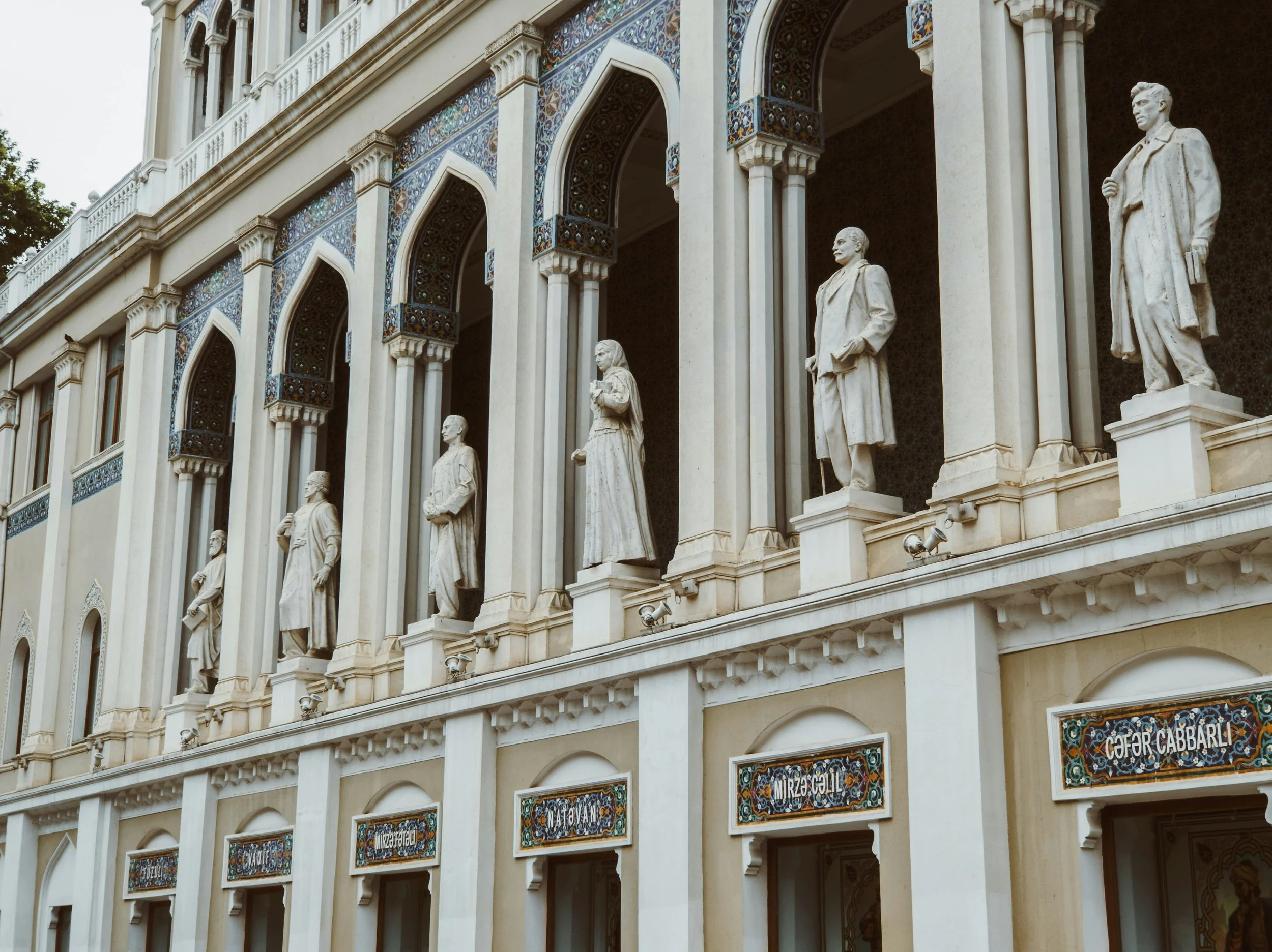 Facade of a building with six white statues of historical figures in niches, decorated with colorful tilework and ornate columns, with rows of plaques below.