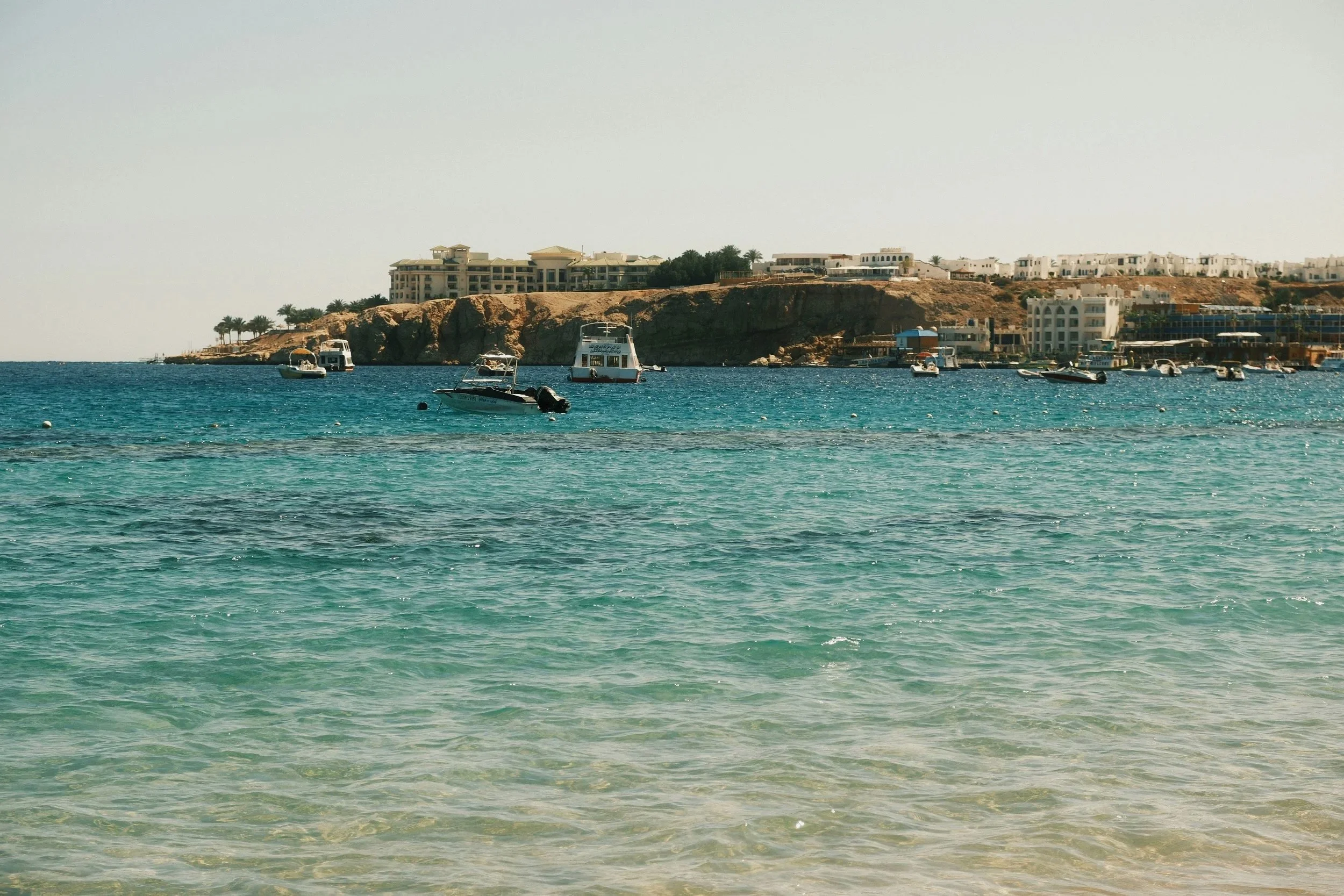 Clear turquoise water with boats floating near a rocky shoreline and buildings on a hill in the background.