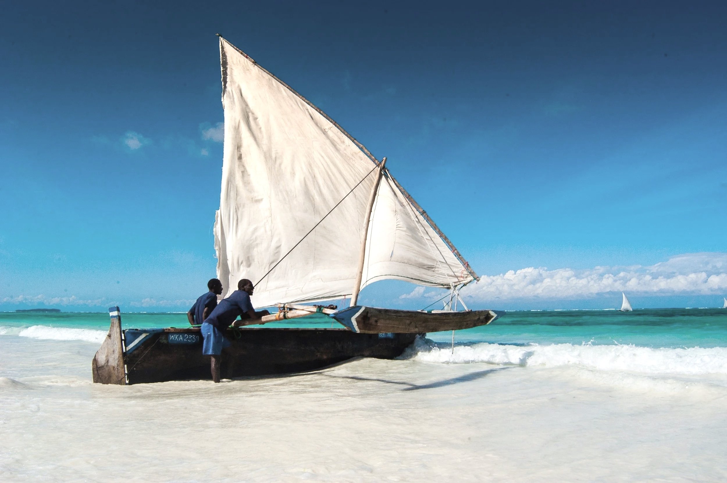 Two boys push a small traditional sailboat onto a sandy beach, with turquoise ocean waves and a clear blue sky in the background.
