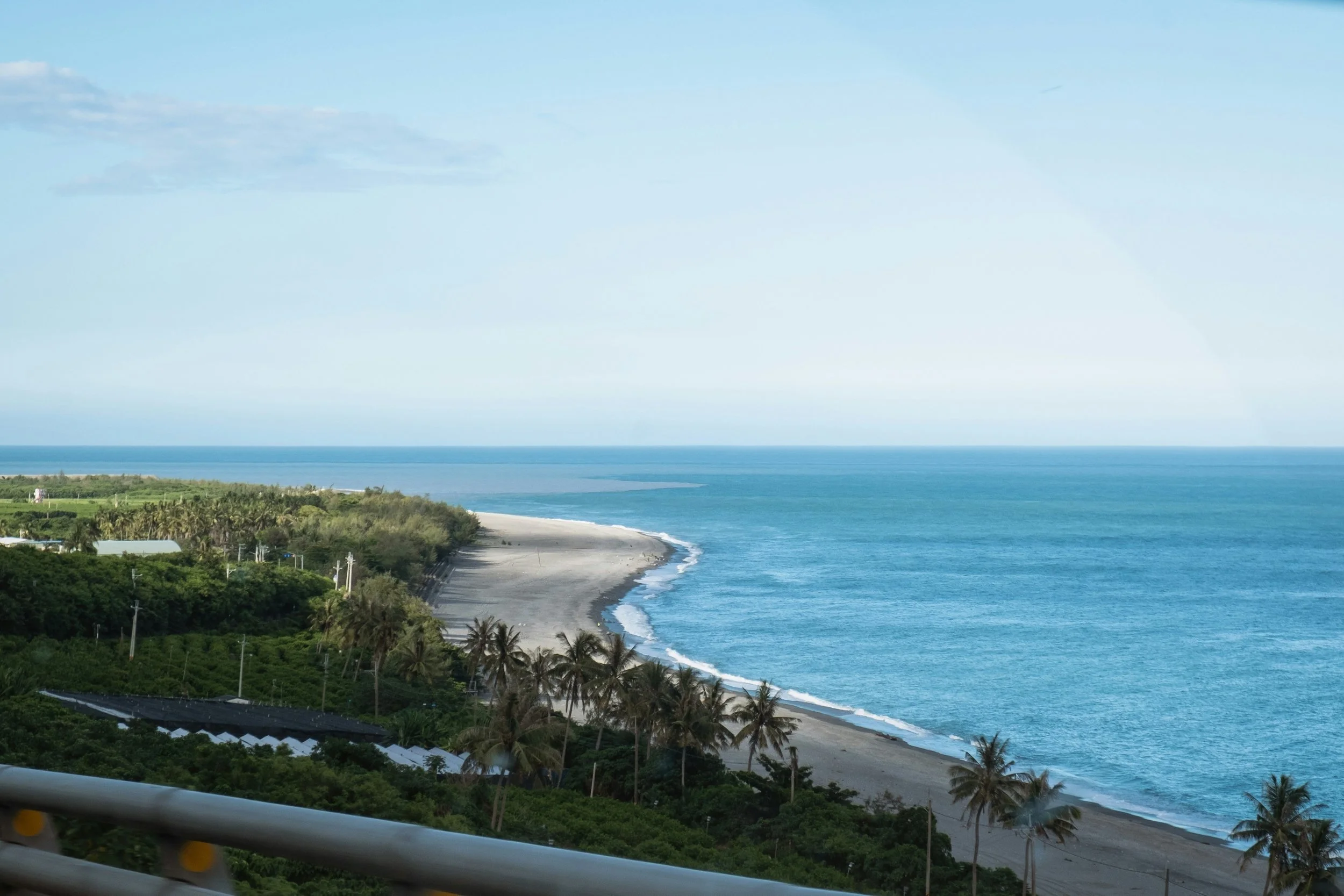Coastal view of a sandy beach, calm blue ocean, and green trees with some palm trees along the shore, under a partly cloudy sky.