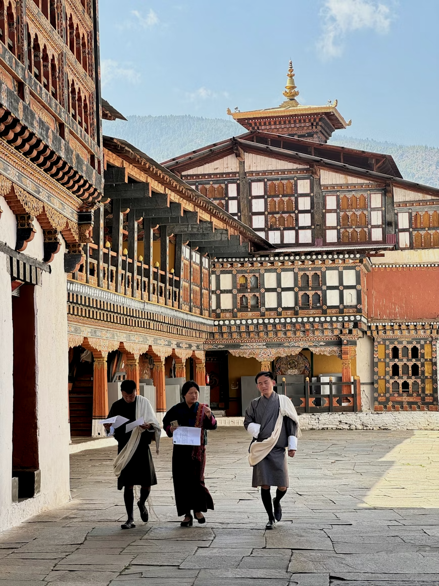 Three Bhutanese monks and a woman walking in a courtyard of a traditional Bhutanese monastery with intricate wooden carvings and layered pagoda-style roof.