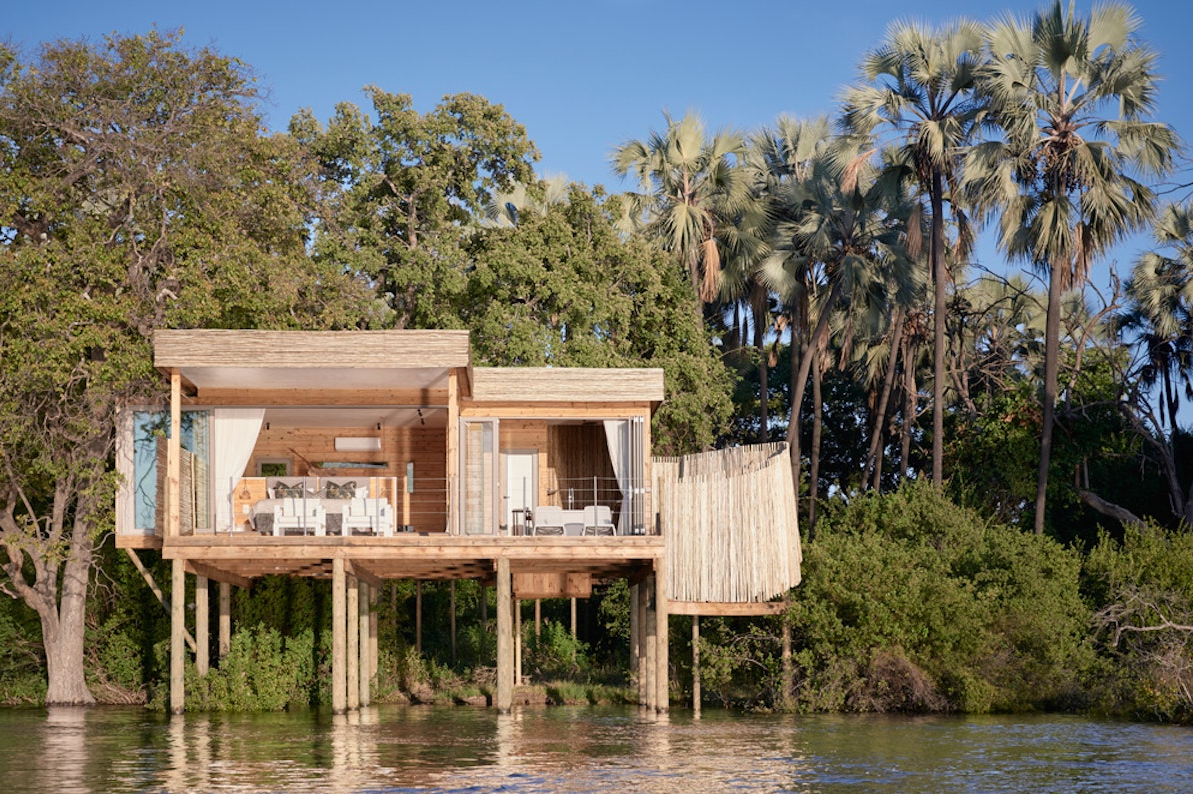 A modern wooden house elevated on stilts over water, surrounded by lush green trees and tall palm trees under a clear blue sky.