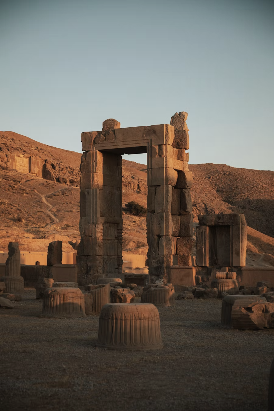 Ancient ruins of stone columns and a rectangular stone arch in a desert landscape at sunset.