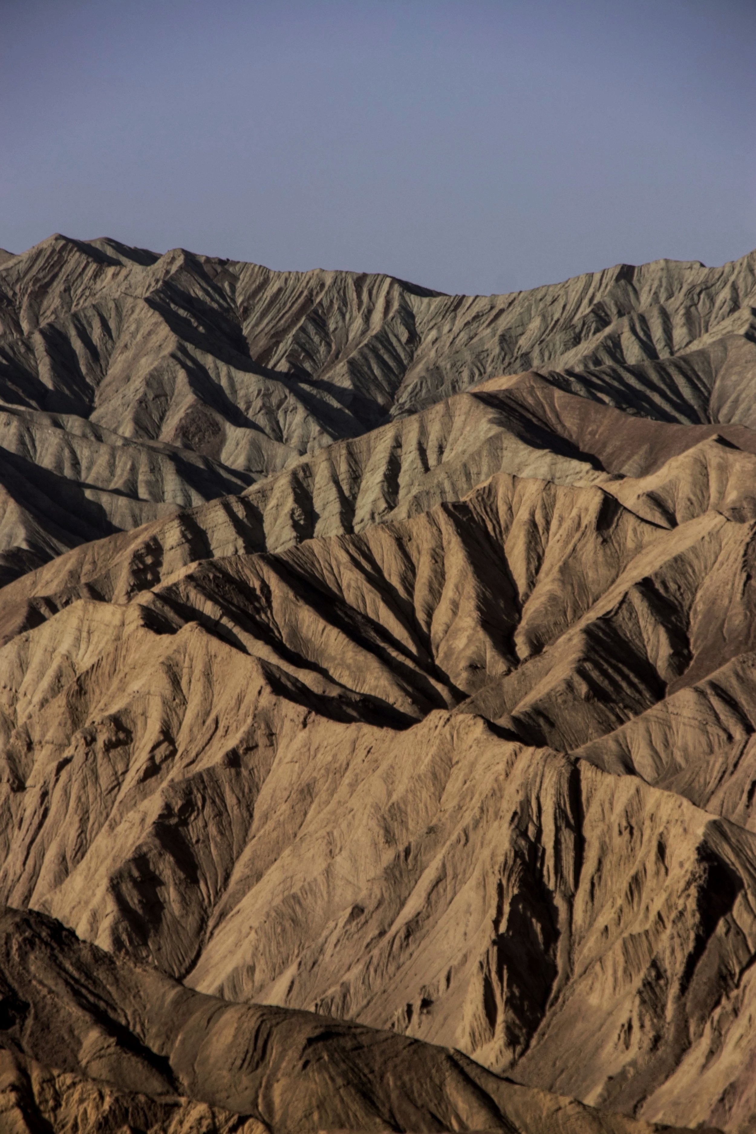 Photo of rugged, barren mountains with deep ridges and layered, multicolored rock formations under a clear, blue sky.
