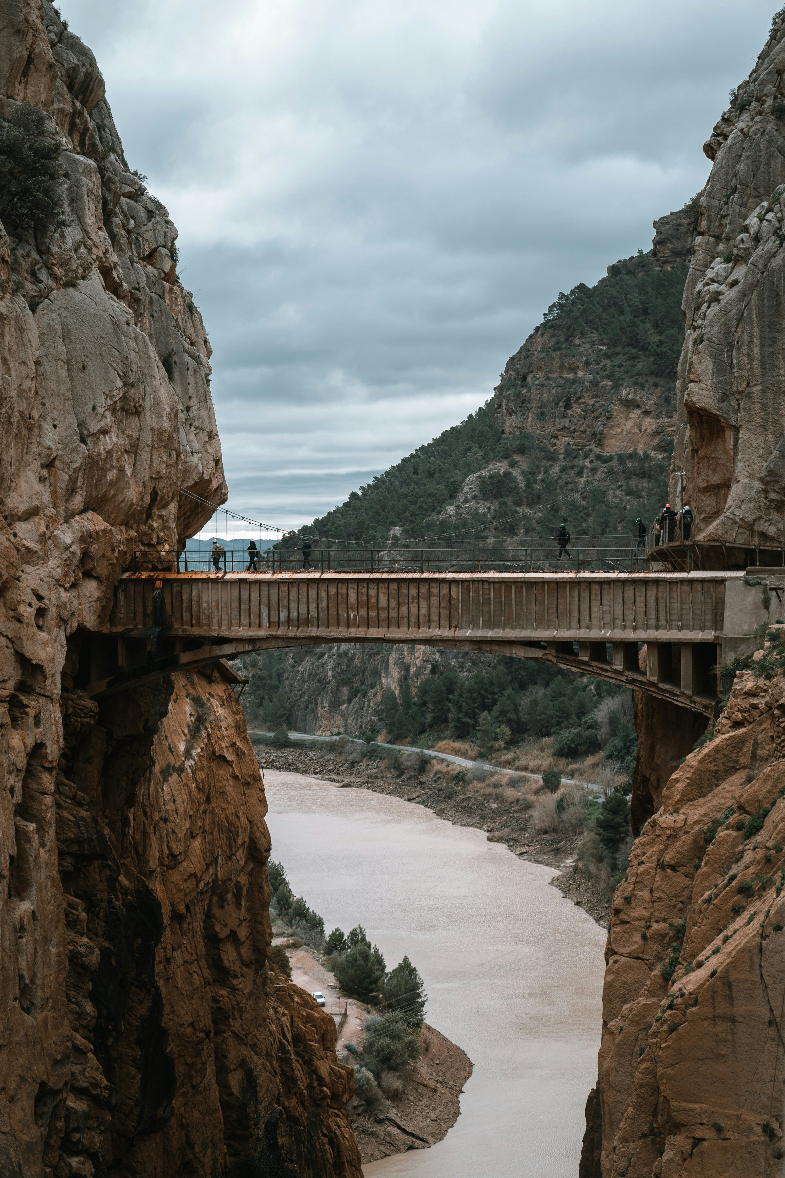 A narrow suspension bridge connects two rocky cliffs over a river in a canyon, with several people walking across it under a cloudy sky.
