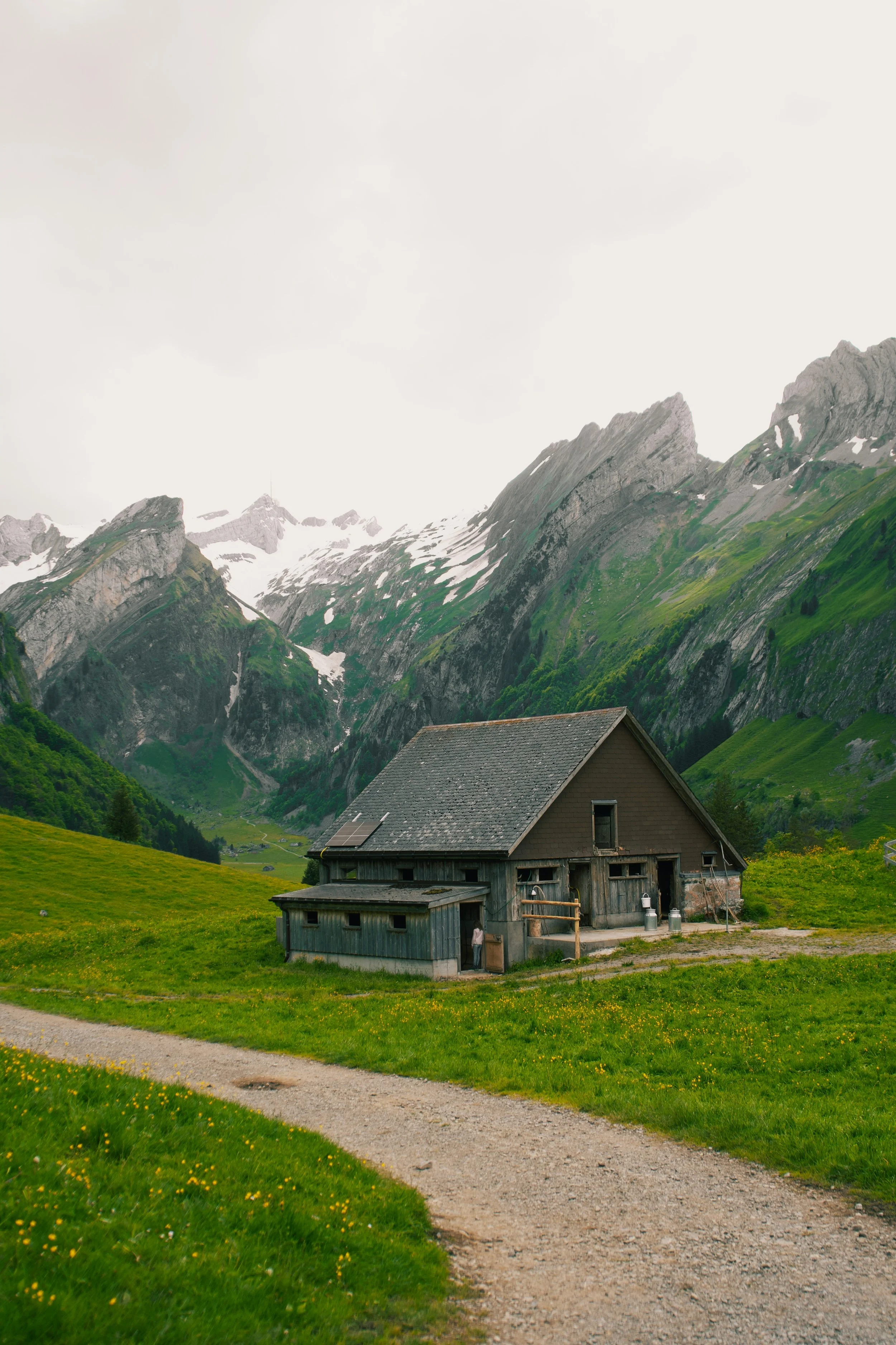 A rustic wooden cabin in a lush green valley with towering mountains in the background and a gravel pathway in the foreground.