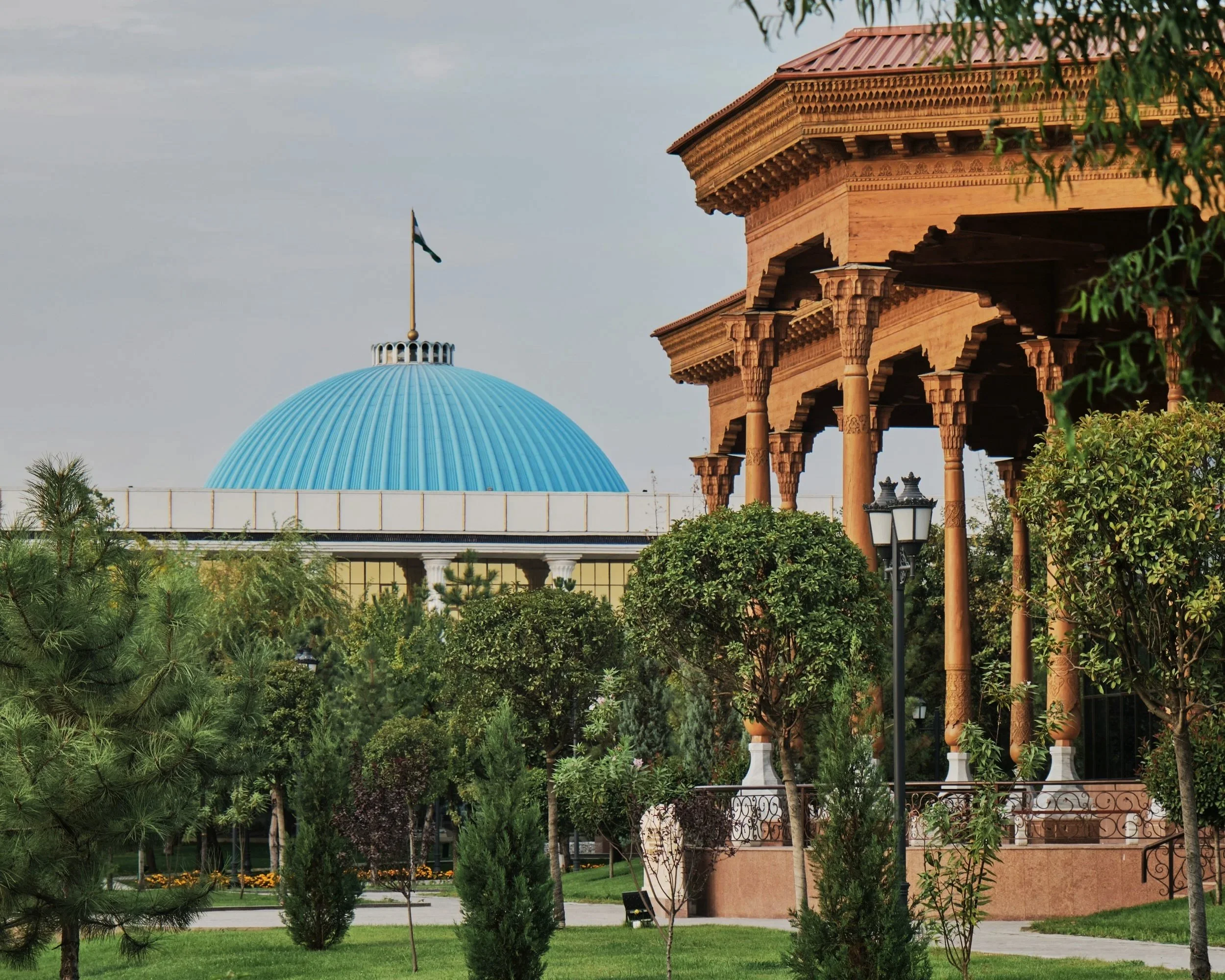 A park with greenery and trees, featuring traditional wooden pavilion with ornate columns on the right, and a large blue-domed building in the background, under a cloudy sky.