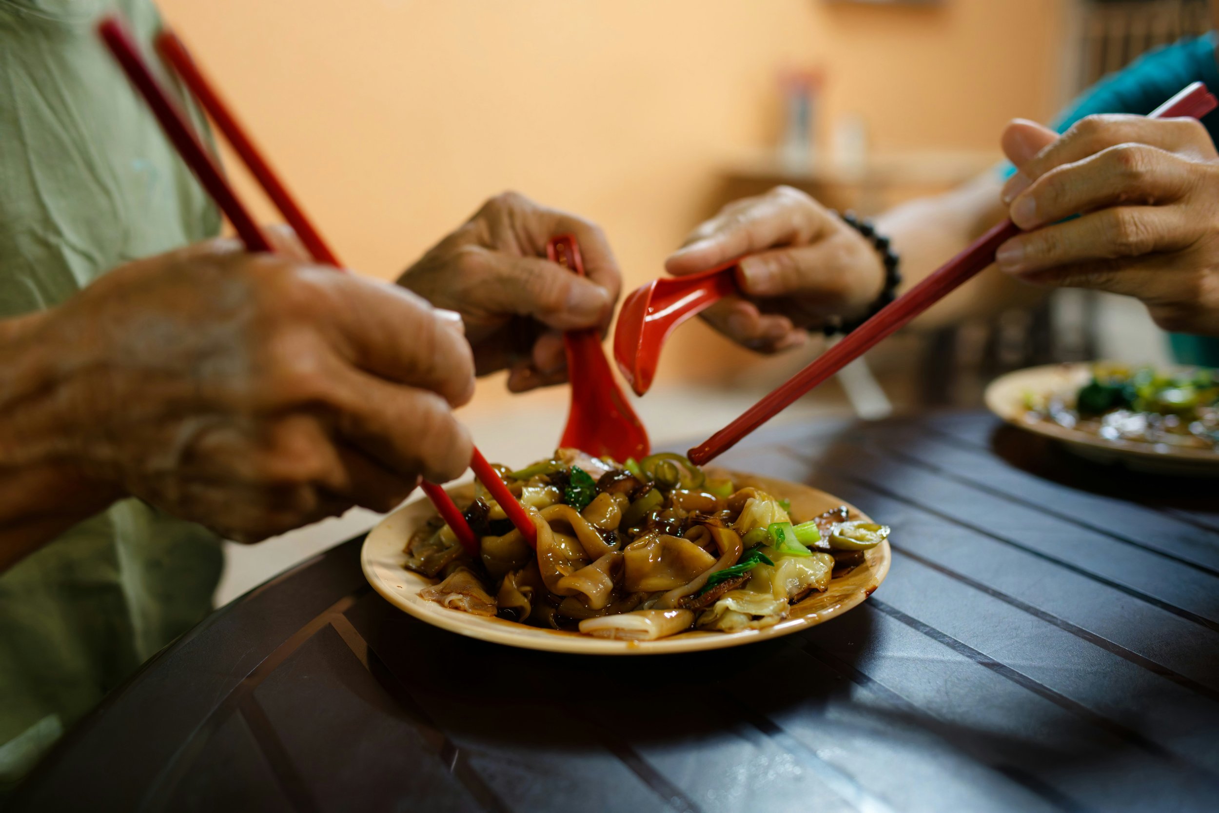 Two people sharing a plate of stir-fried noodles with vegetables, using chopsticks and a serving spoon.