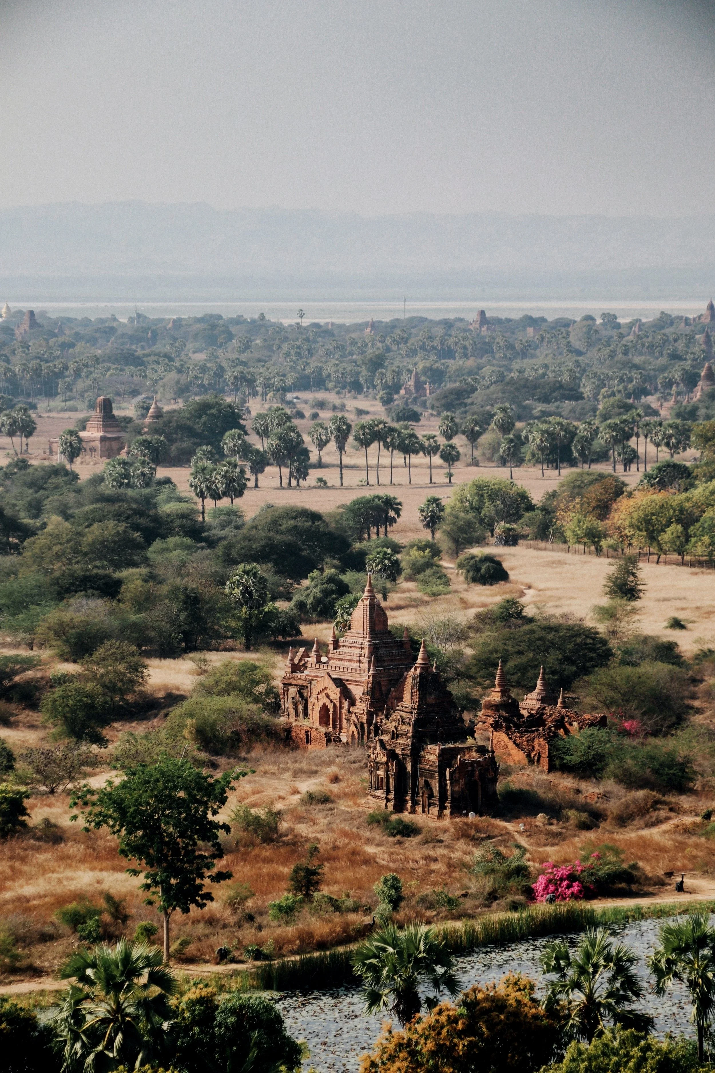 Ancient temple ruins surrounded by trees and vegetation, with a body of water and distant mountains in the background.