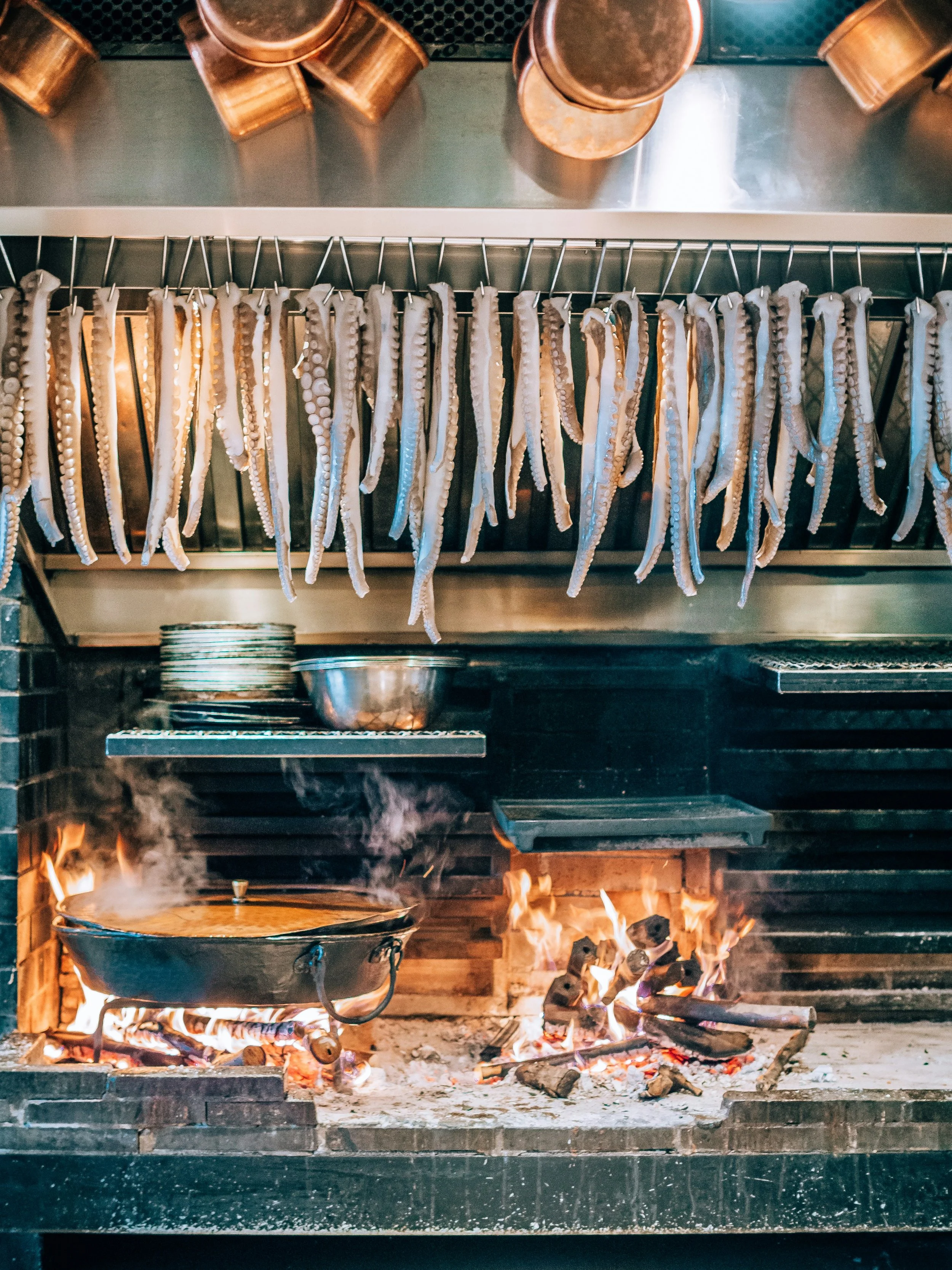 Octopus tentacles hanging above a large open wood fire cooking in a pan.