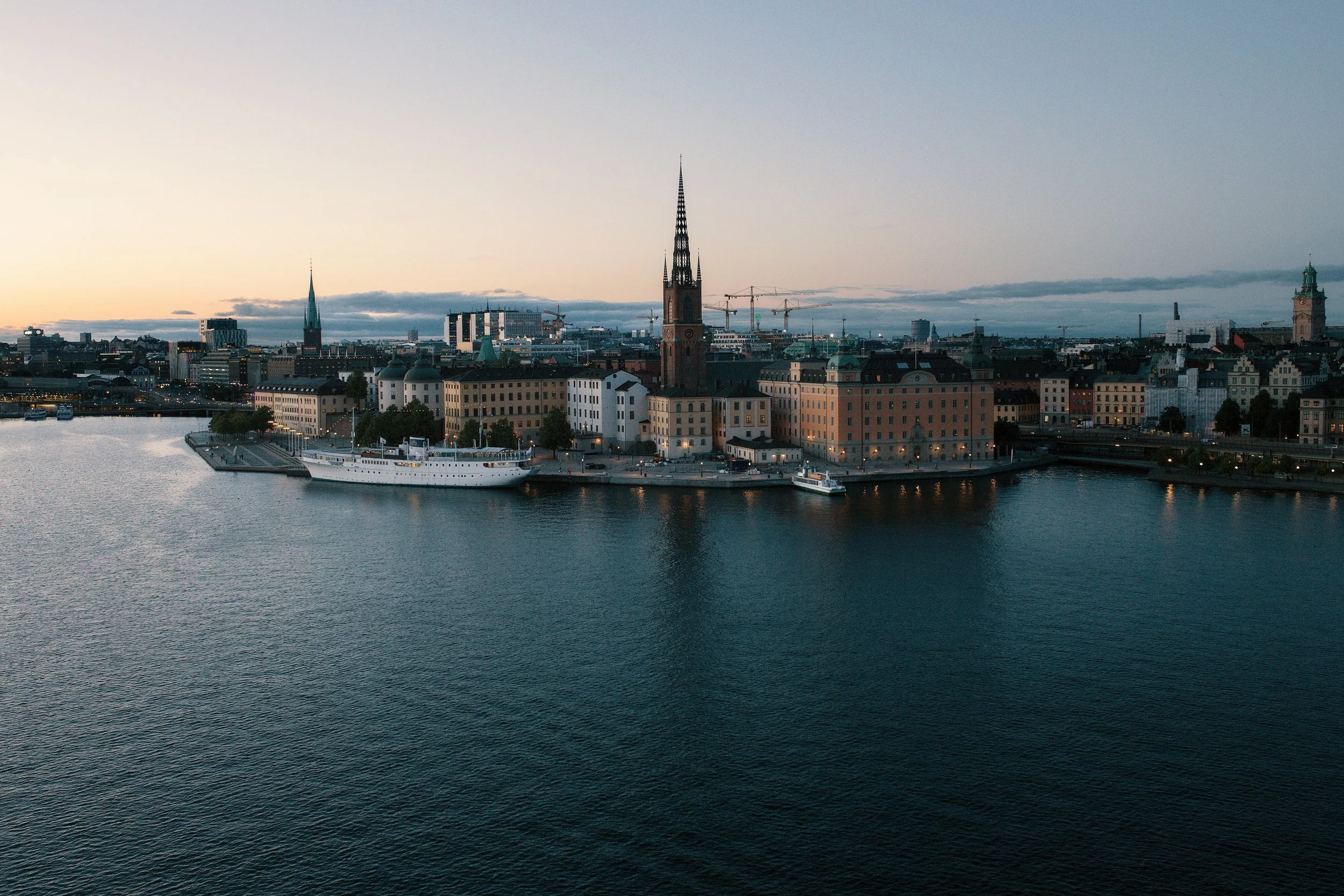 City skyline at dusk with historic buildings, including a church with a tall spire, alongside a body of water with boats.