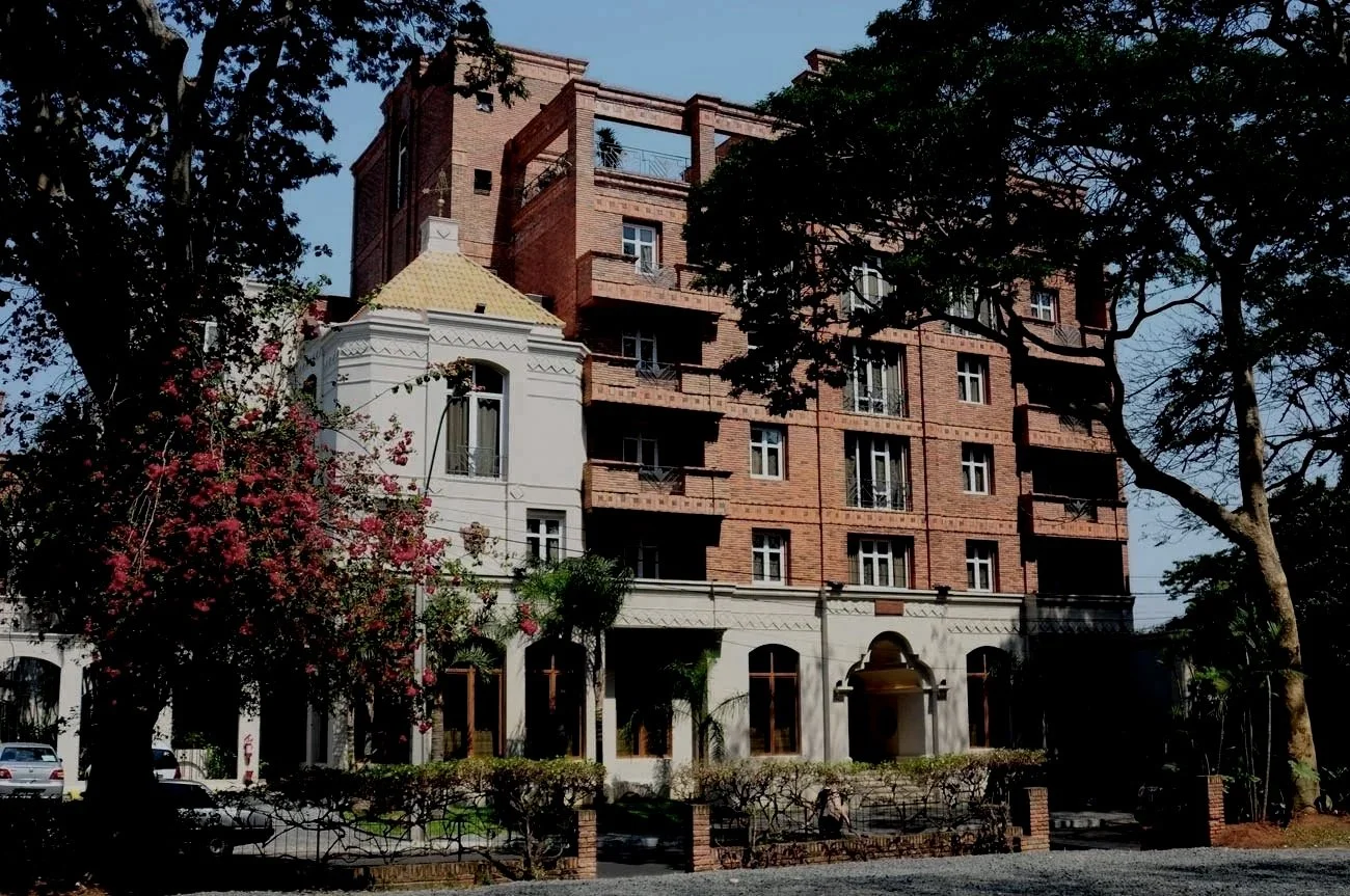 A multi-story building with a mix of brick and white exterior walls, surrounded by trees and a small parking area in front.