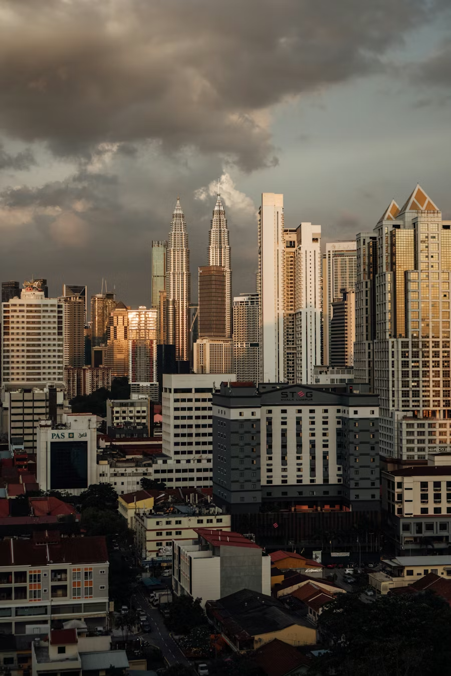 City skyline featuring the Petronas Towers and various modern high-rise buildings during sunset with dark clouds overhead.