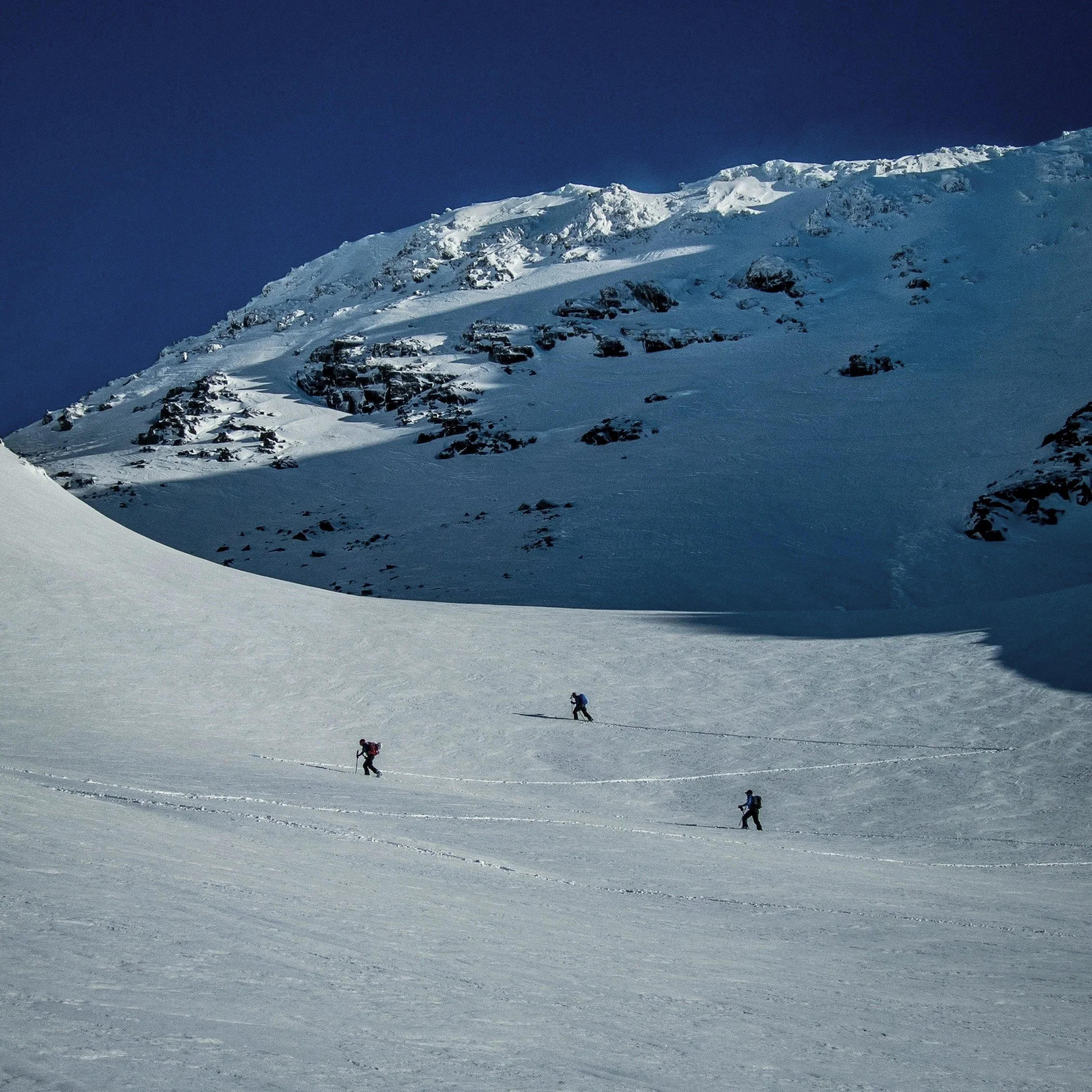 Three people skiing on a snow-covered mountain slope with peaks and rocky outcroppings in the background under a clear blue sky.