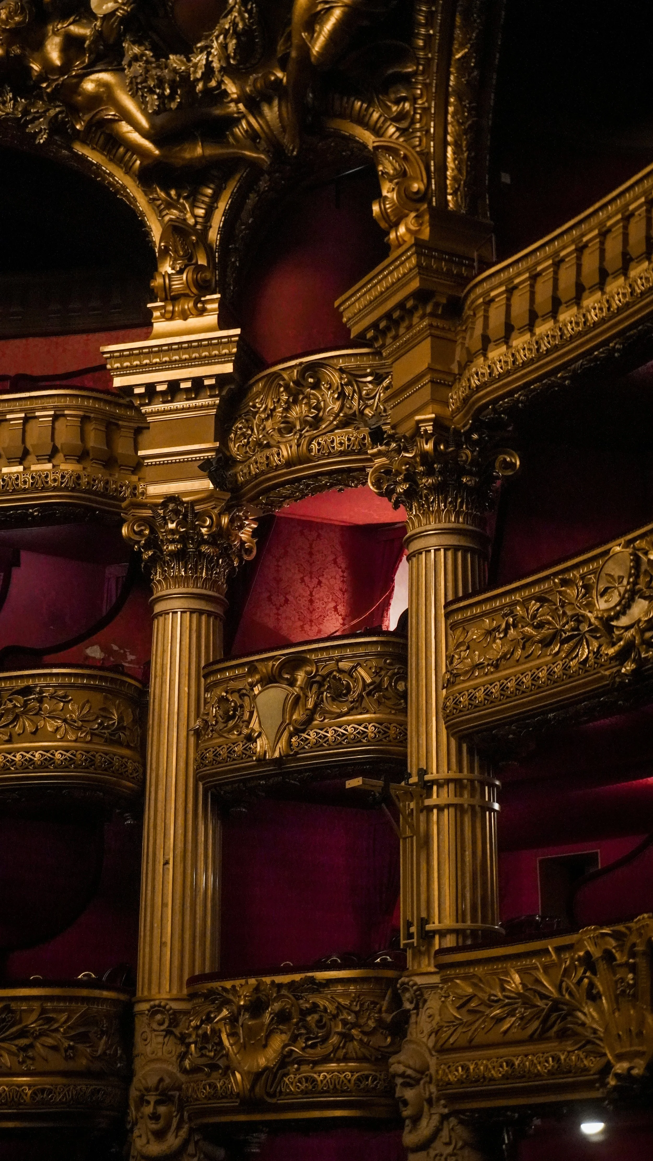 Ornate theater balcony with gold decorative details, columns, and red velvet drapes.