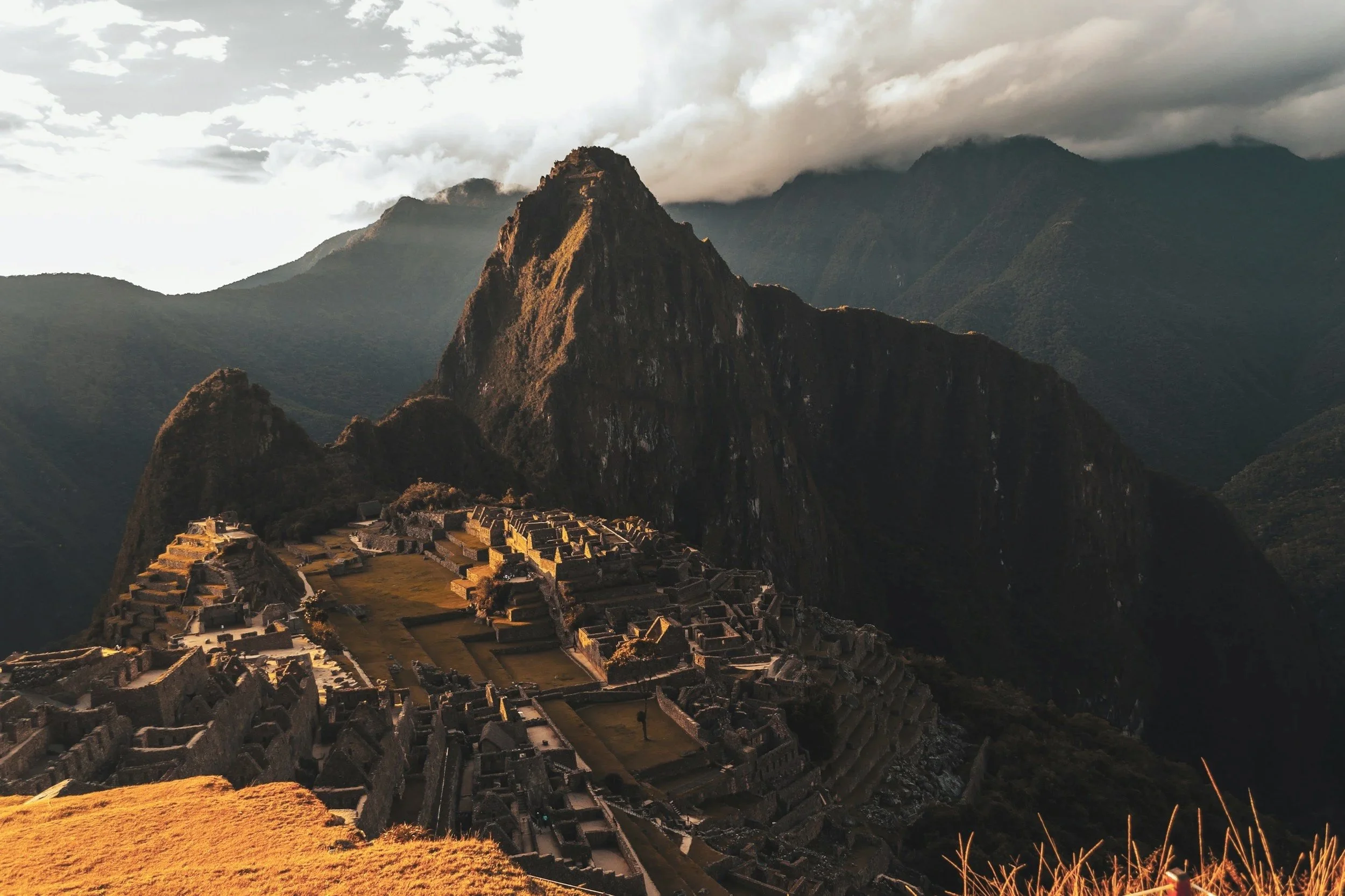 Sunlit view of Machu Picchu with ancient ruins and terraced fields at the base of a steep mountain, with cloudy sky in the background.