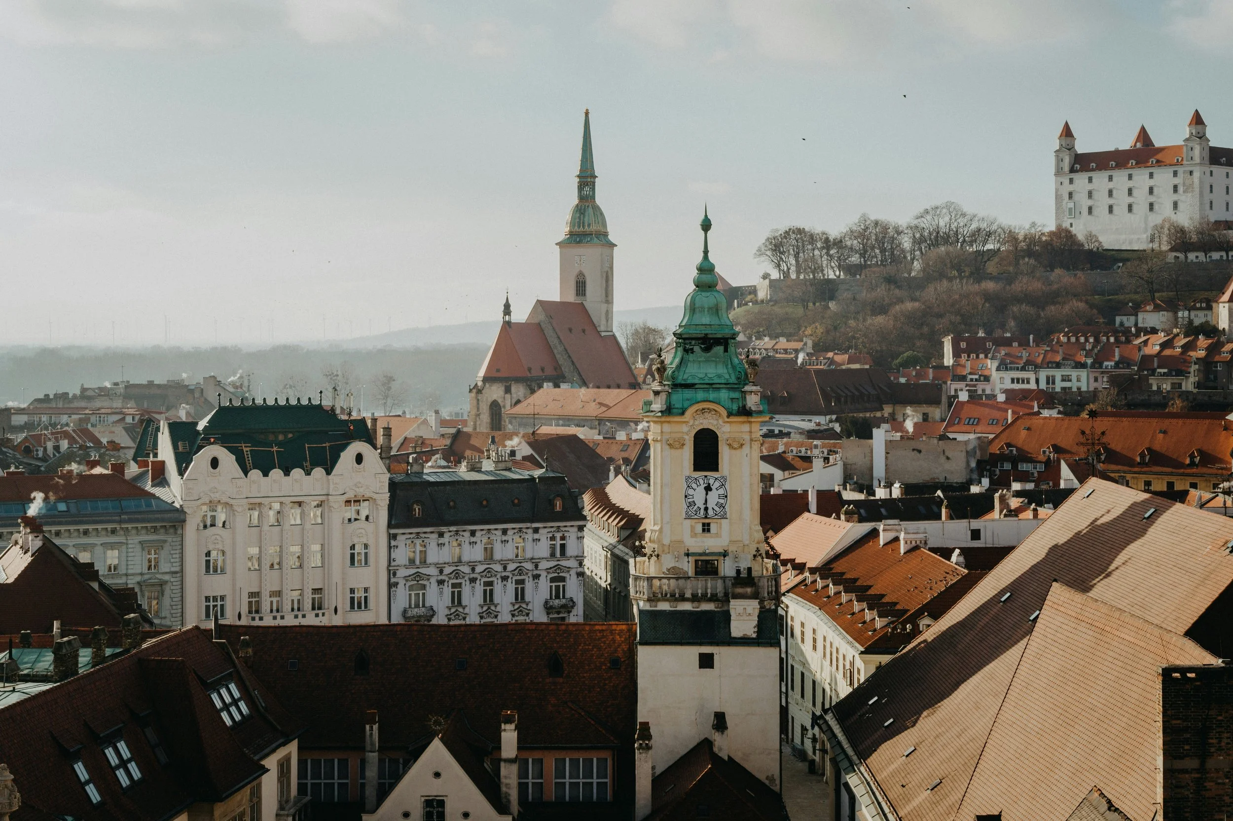 A cityscape view showcasing historic buildings with red-tiled roofs, a tall clock tower with a green spire, and a castle on a hill in the background under a clear sky.
