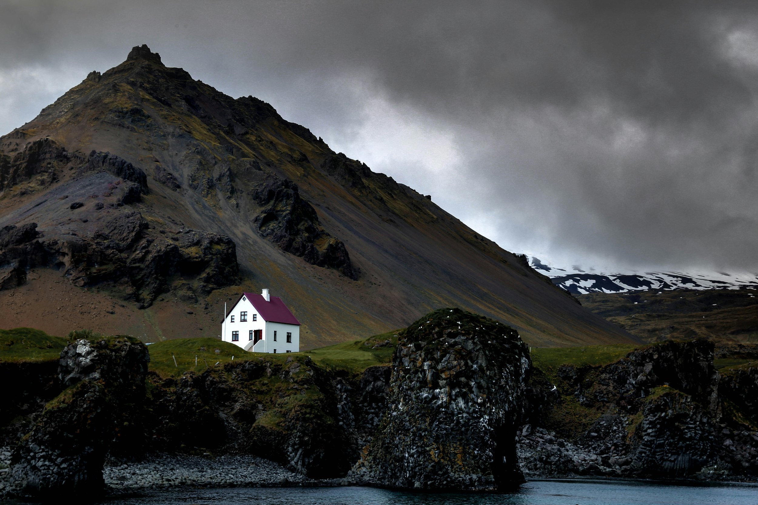 A white house with a red roof sits on a grassy hill by the water's edge, with rocky cliffs and a mountain with patches of snow in the background under a dark, cloudy sky.