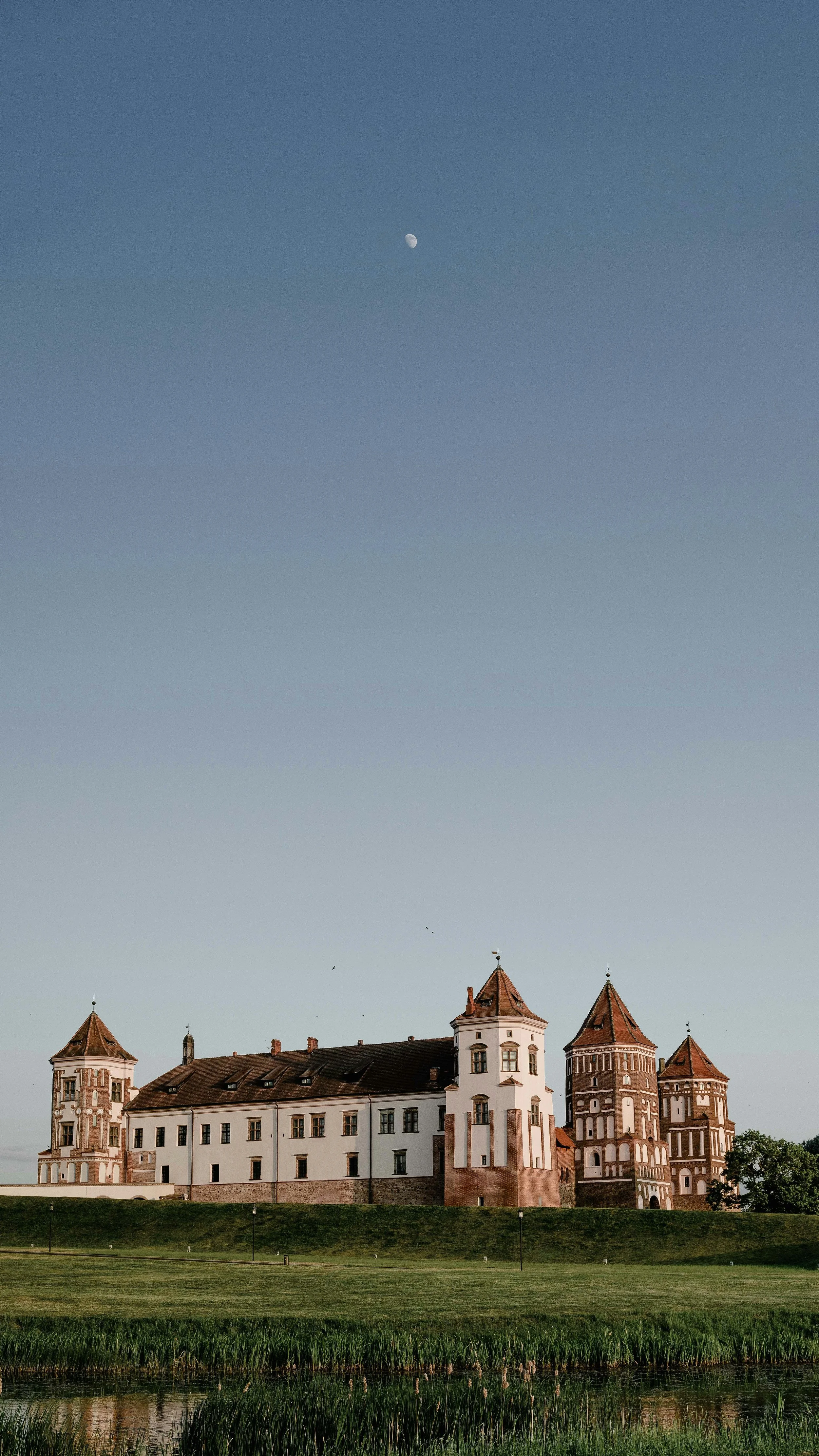 A historic castle with multiple towers and white walls, surrounded by green grass and trees. The sky is clear, with a visible moon and a few birds flying.