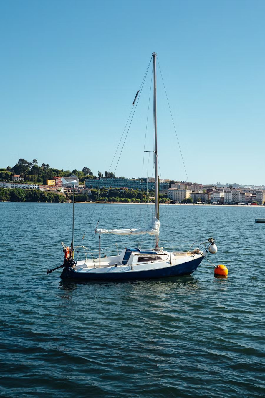 A sailboat floating on calm water near a buoy with a cityscape and green hills in the background.