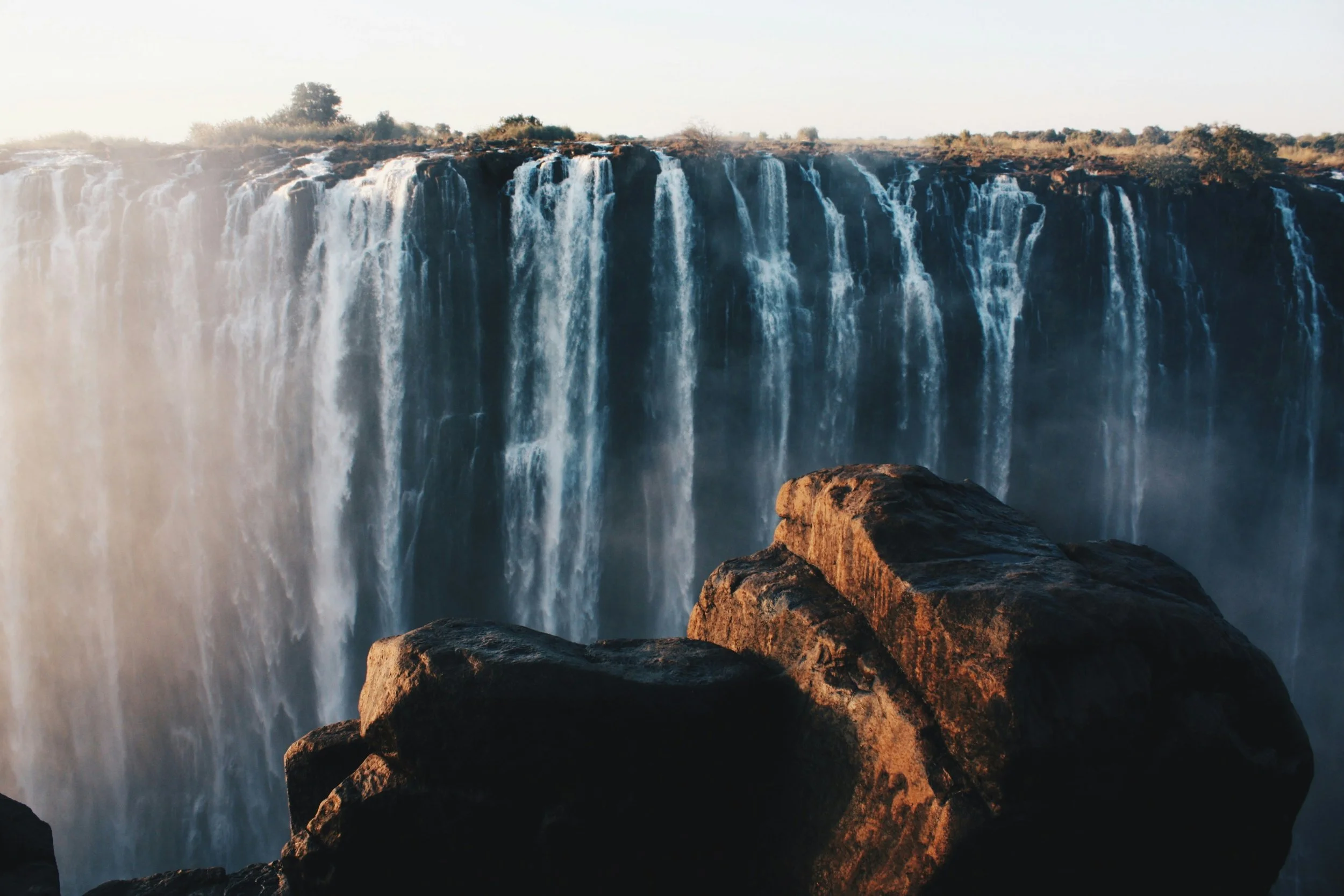 Waterfalls cascading over a cliff with rocks in the foreground during sunset.
