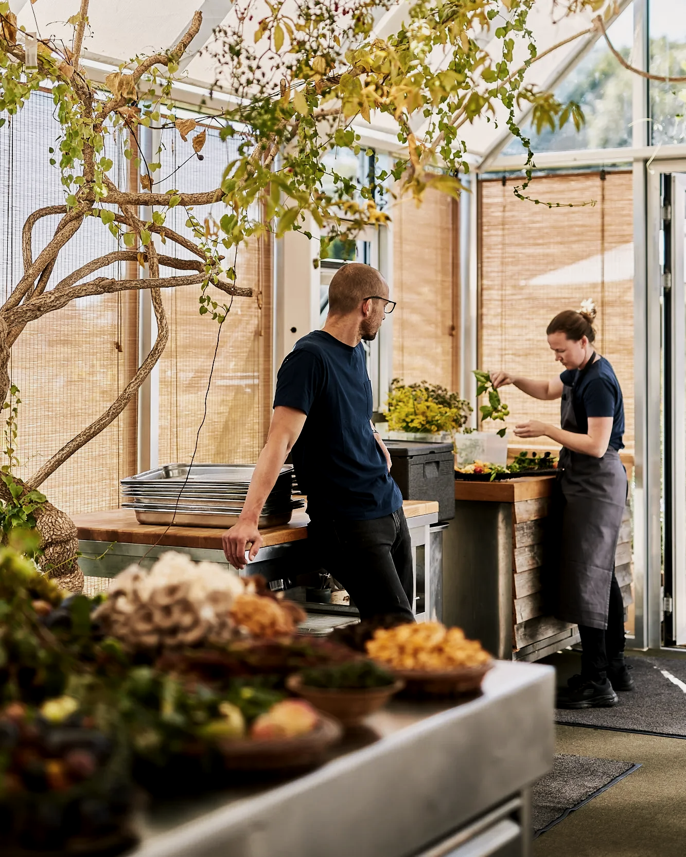 Two people working in a greenhouse with plants and wooden decor, one leaning on a table and another picking leaves.