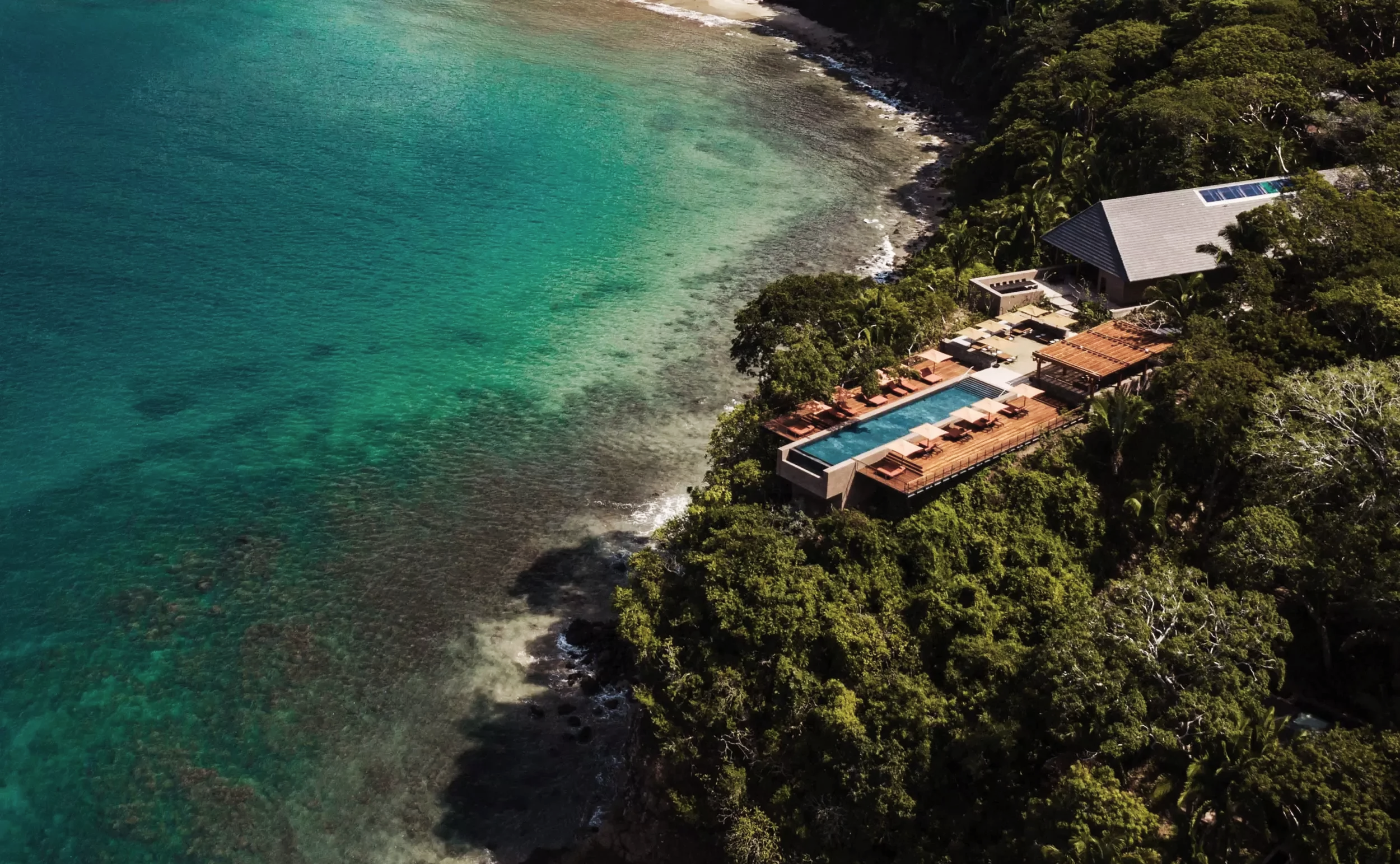 Aerial view of a luxury seaside property with a rooftop pool, surrounded by lush greenery and situated along a clear, turquoise coast.