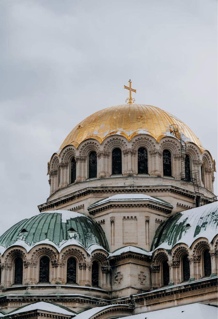 Close-up of a church with three domes, the central one gold with a cross on top, and the two green domes on either side, all with snow on top, against a cloudy sky.