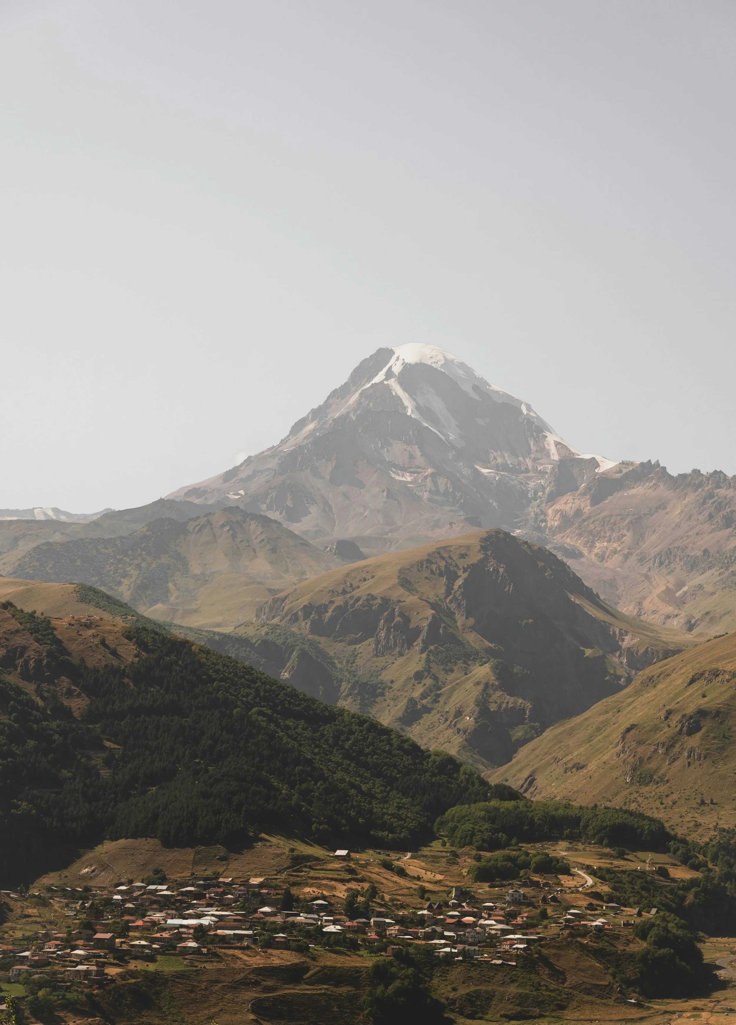 A mountain with snowy peak surrounded by green hills and a small village in the foreground.
