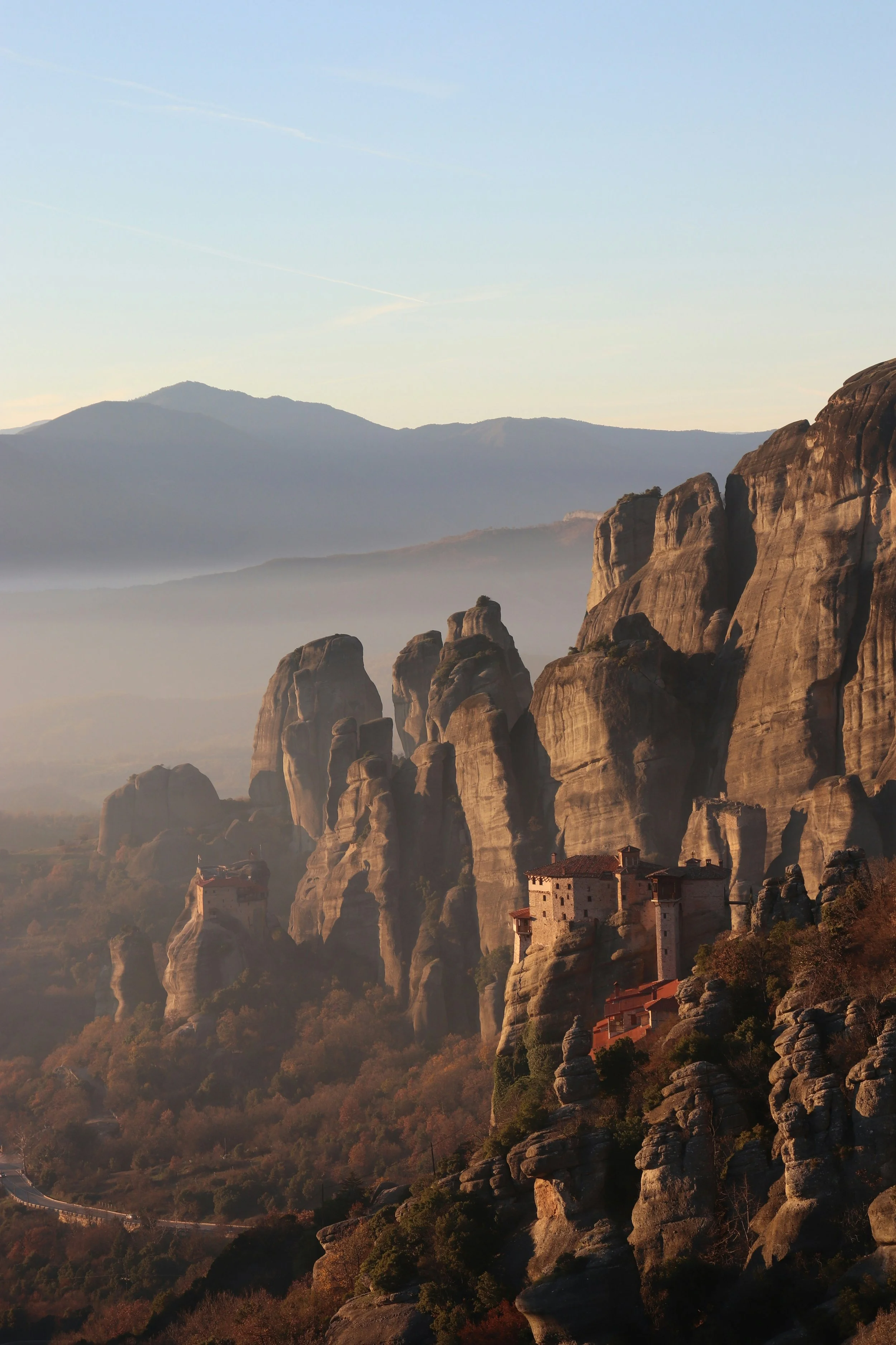 Mountains with rocky formations and a monastery built into the cliffs, with a hazy sky in the background during sunset.