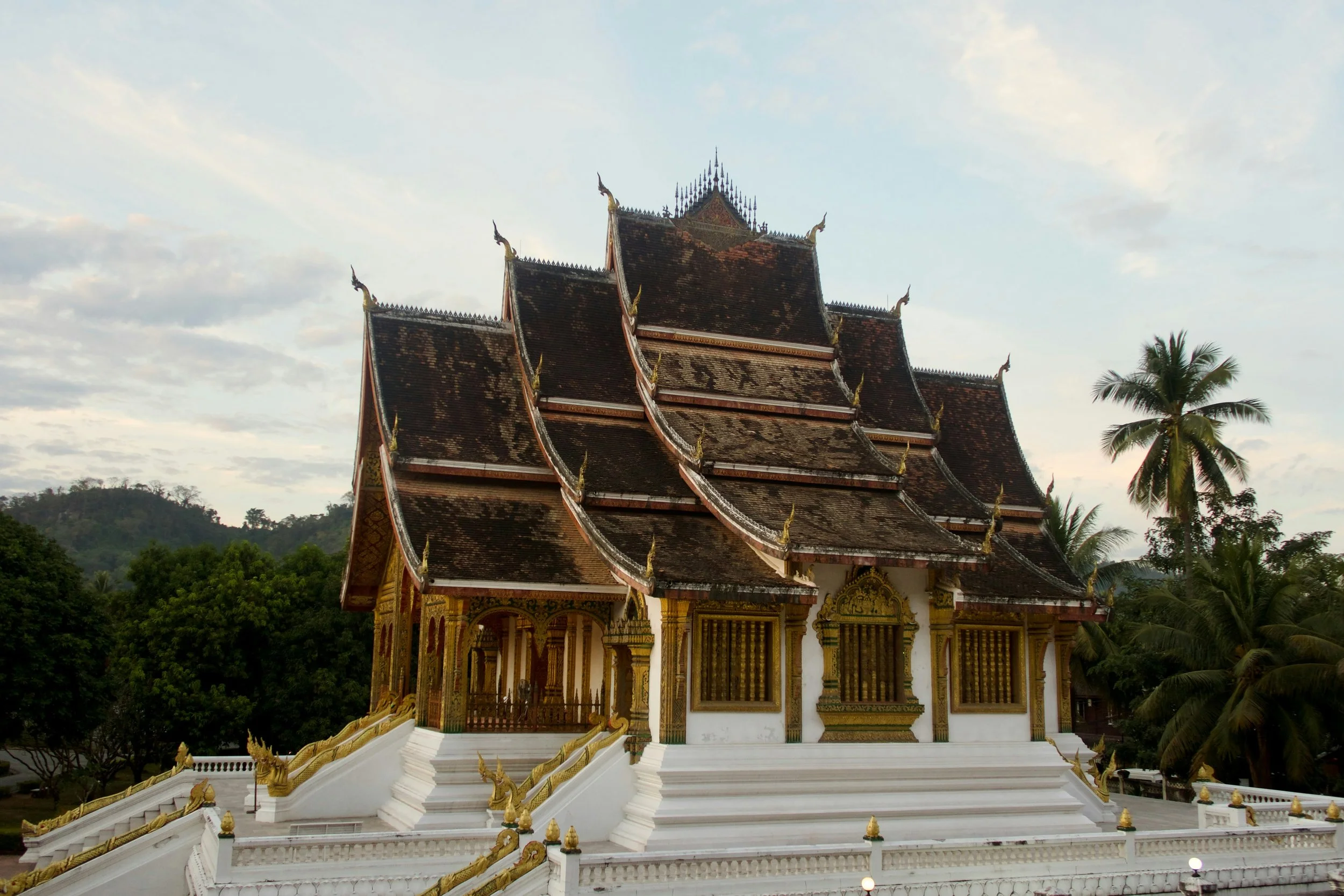 Traditional multi-tiered Thai temple with ornate gold decorations, white stairs, and dark, weathered roof, surrounded by palm trees and greenery.