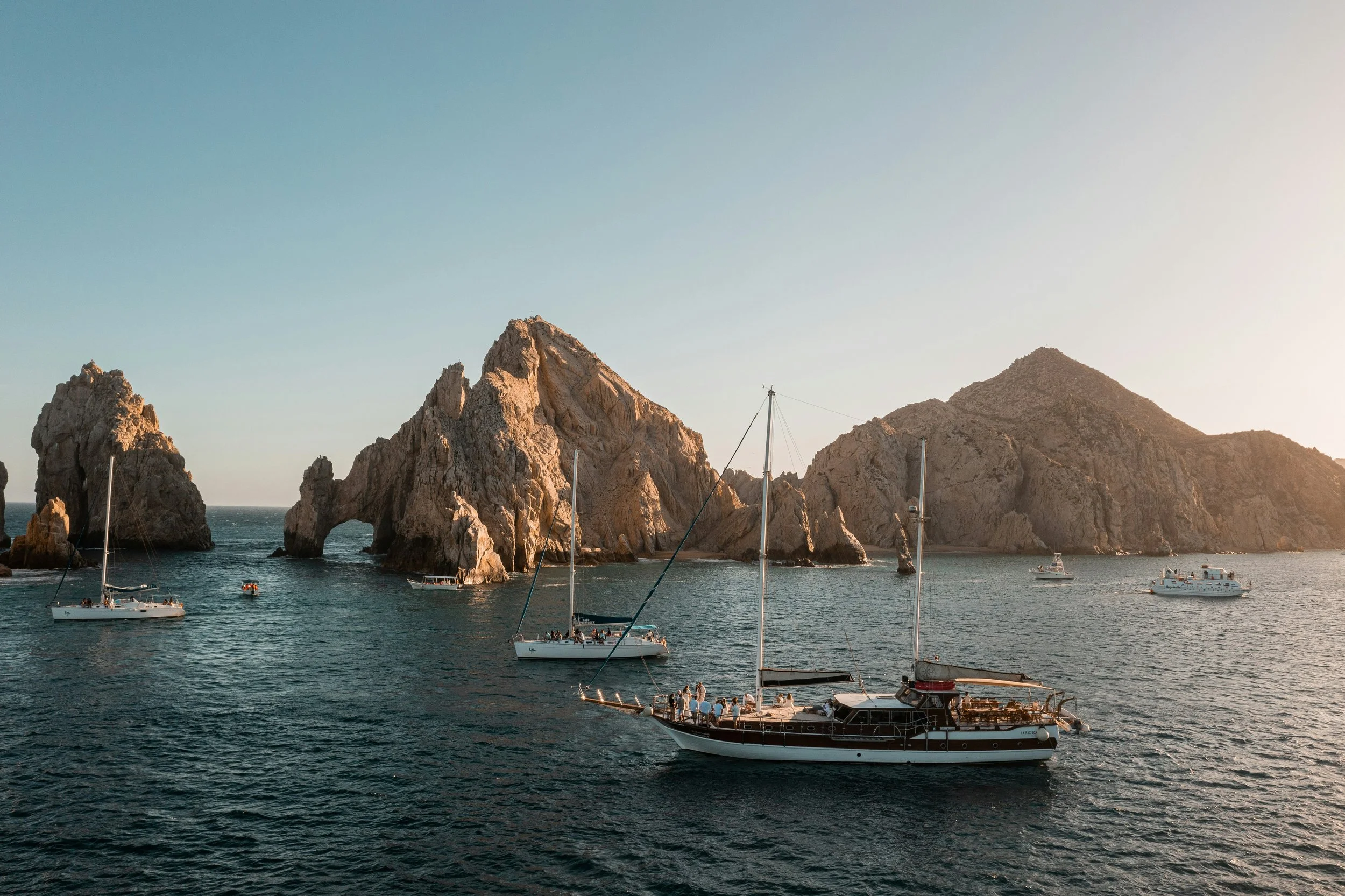 Sailboats anchored near a rocky coastline during sunset.