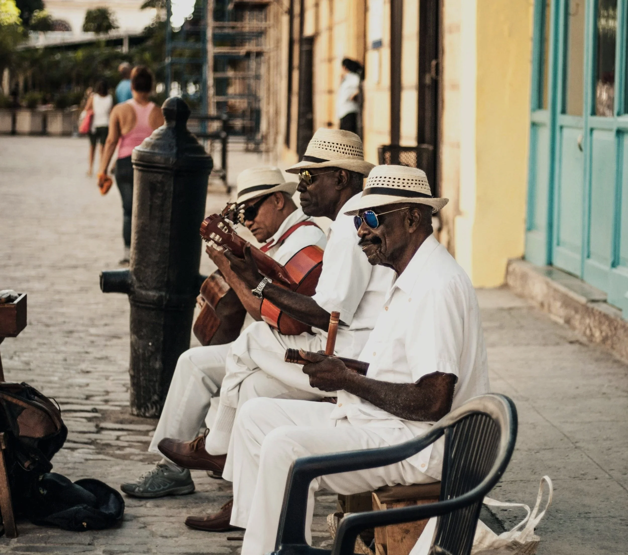 Three elderly men dressed in white, wearing straw hats and sunglasses, sitting on the sidewalk playing musical instruments in a street scene.