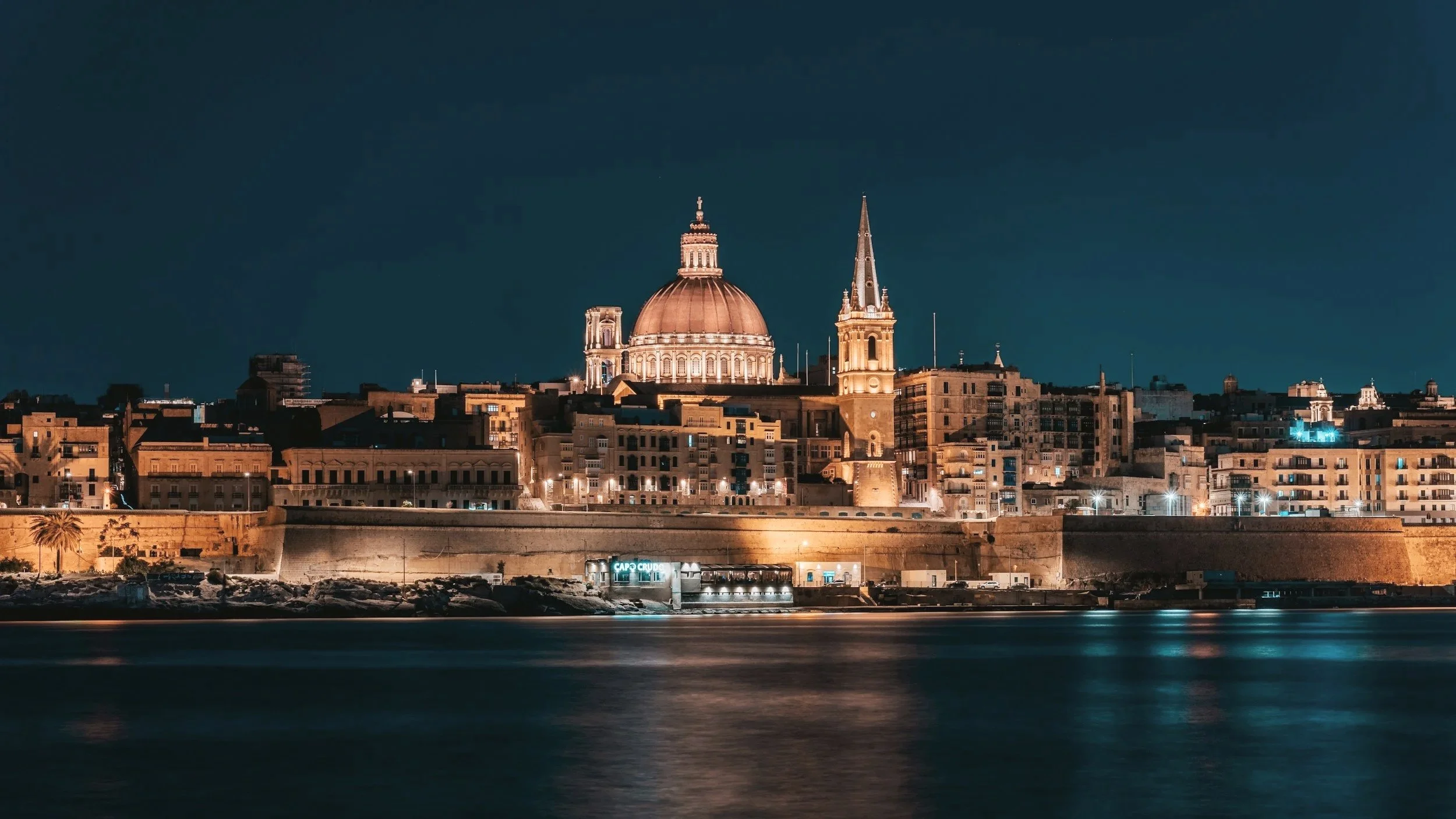 Nighttime view of a cityscape with historic buildings, including a prominent domed church and tall spire, illuminated and reflected in the water in the foreground.